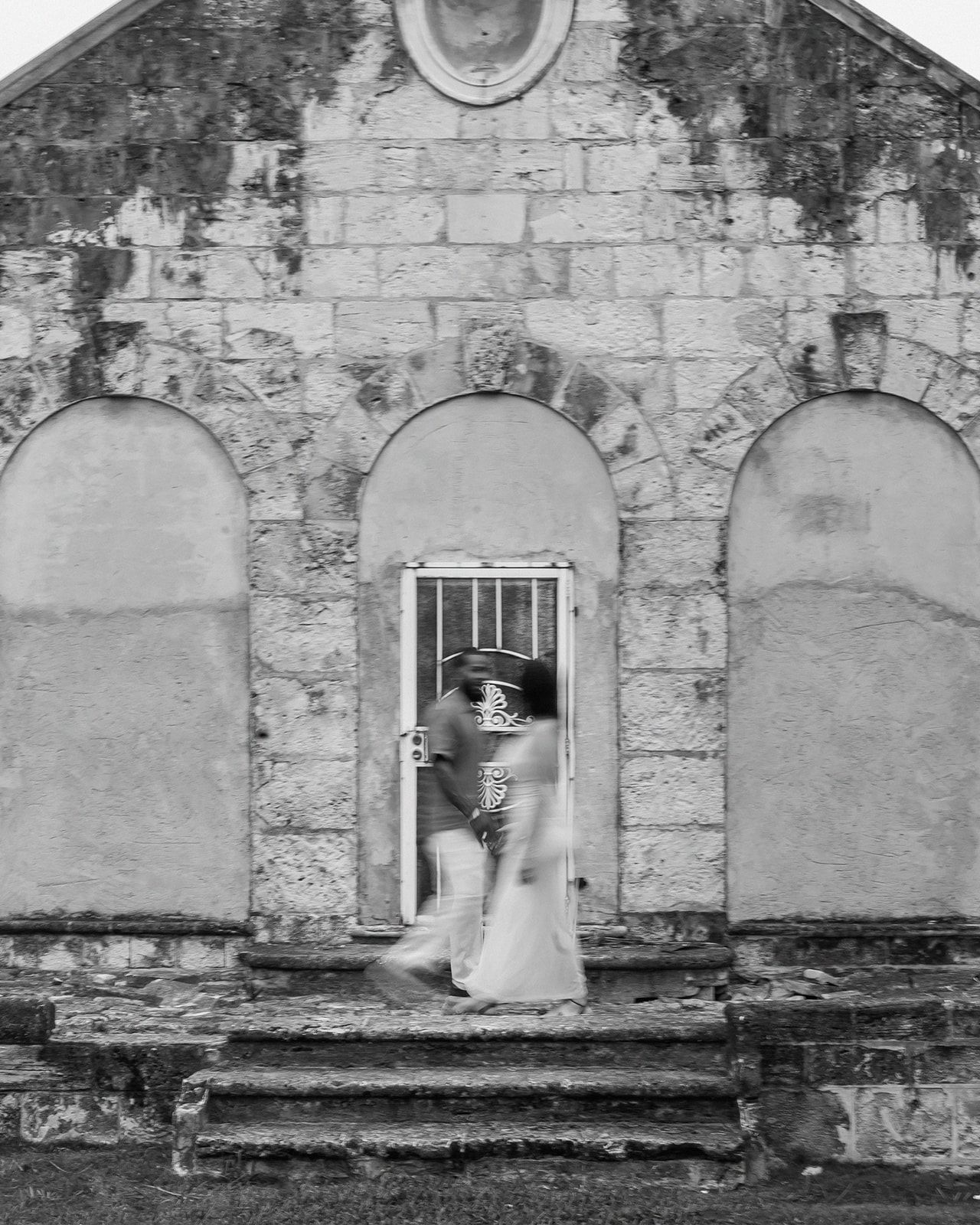 Black and white photo of a couple kissing in a doorway of a rustic stone chapel on their wedding day.