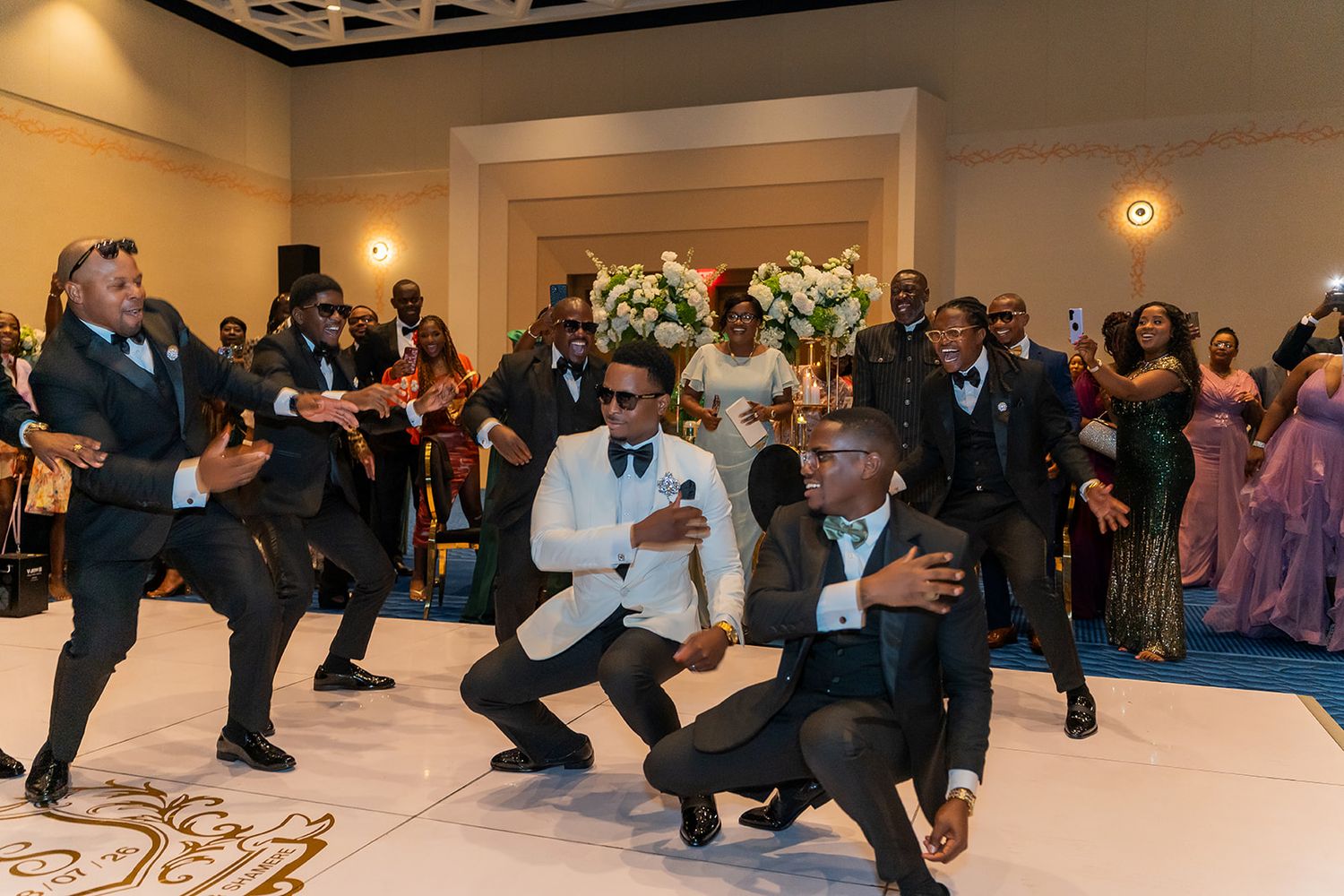 Groomsmen performing an energetic dance routine at a wedding reception in an elegant ballroom with guests watching.