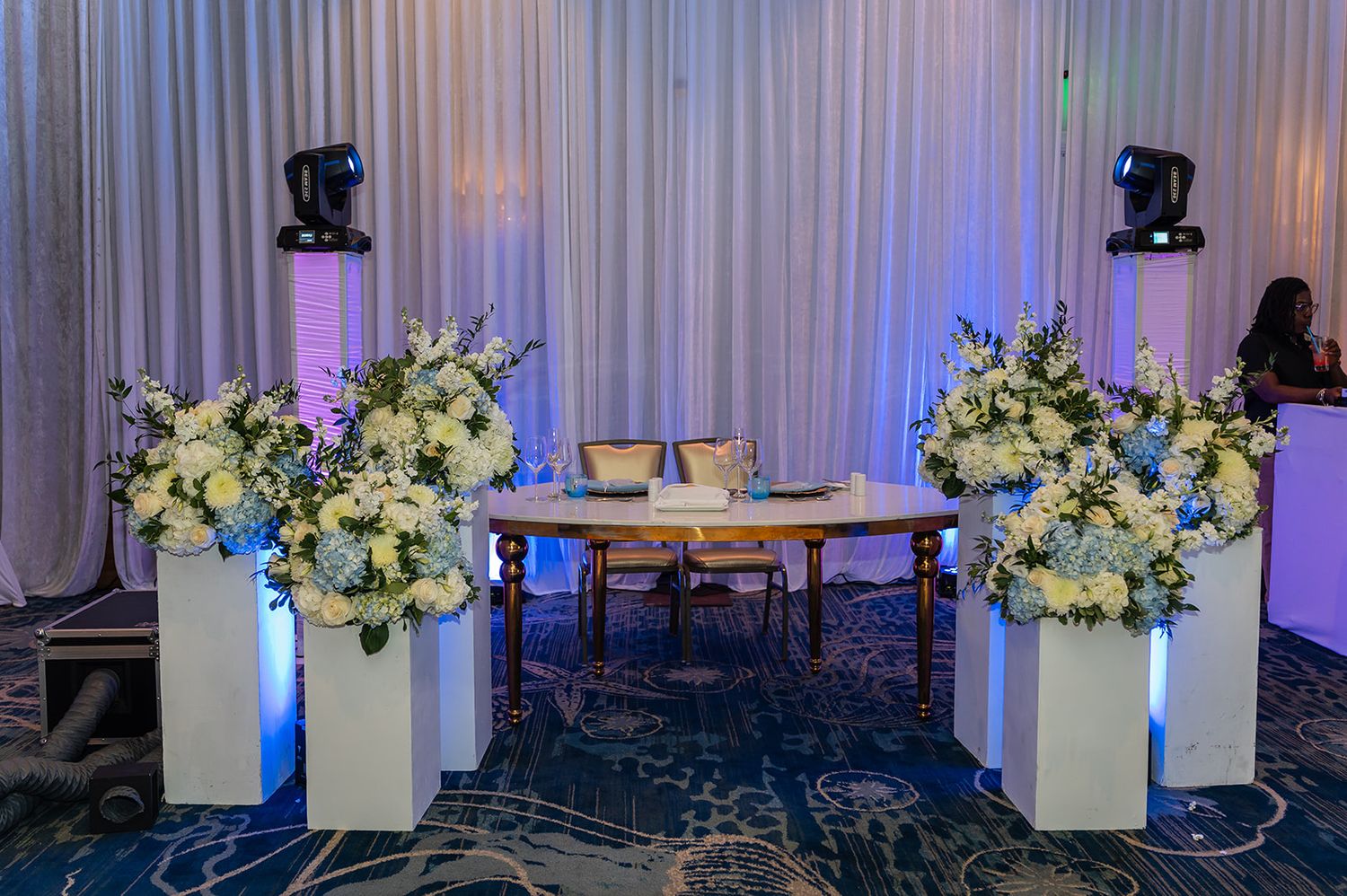 Elegant wedding sweetheart table with white floral arrangements on pedestals and blue uplighting in a ballroom.