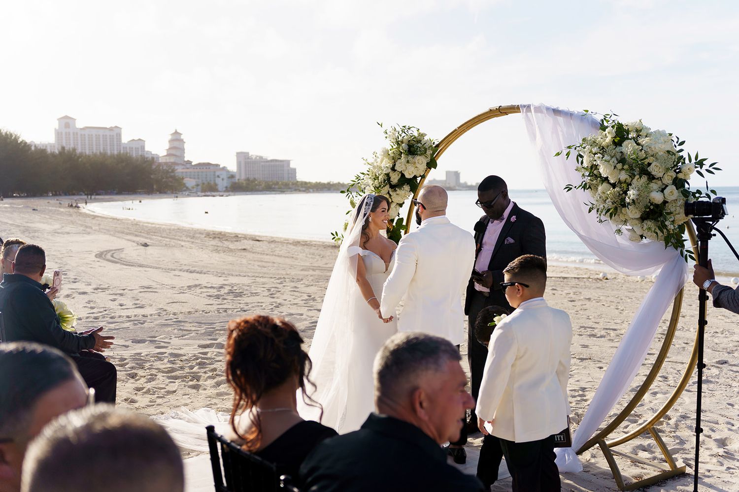 Couple exchanges vows at a romantic beach wedding ceremony under a gold circular arch adorned with white flowers.
