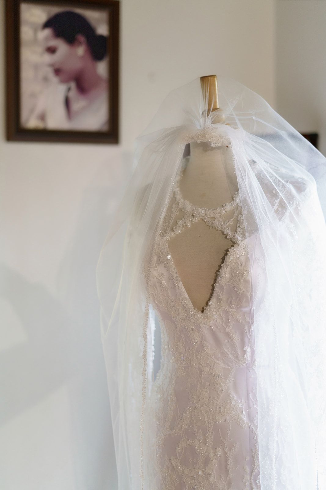 Elegant lace wedding dress with veil on mannequin, keyhole back detail, framed photo in background.