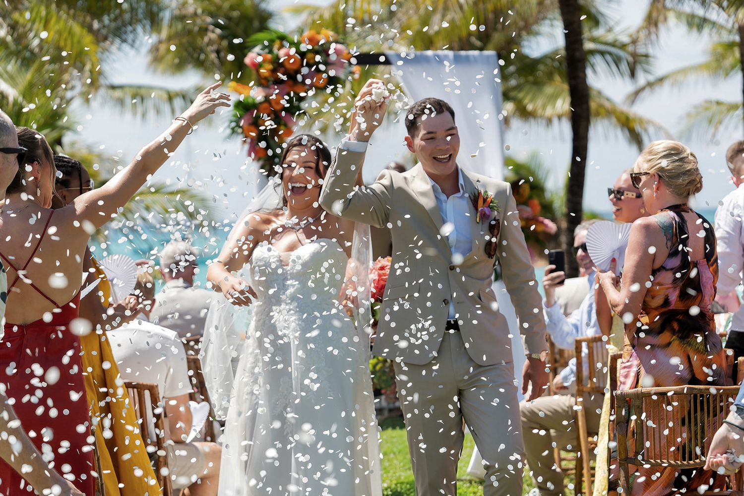 Joyful newlyweds walk through confetti shower outdoors at tropical beach wedding ceremony with palm trees.
