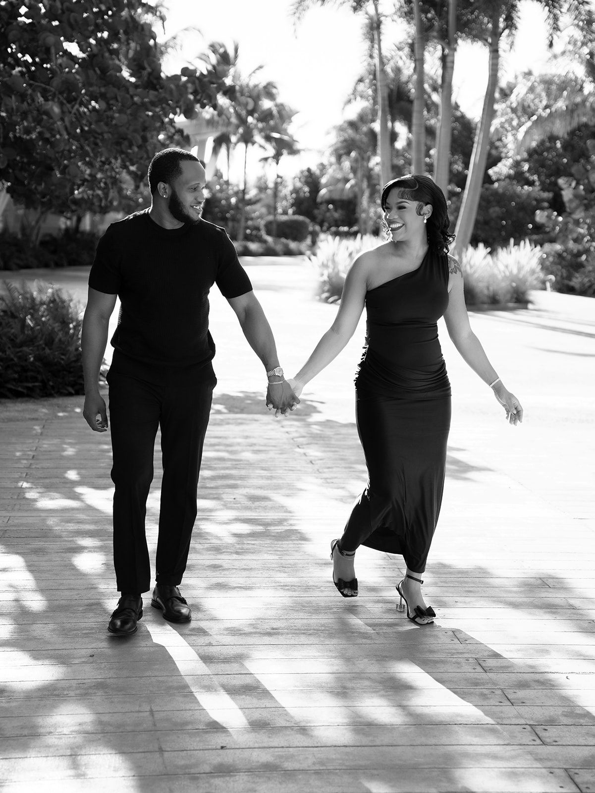 Black and white photo of smiling couple holding hands and walking along a palm tree-lined pathway.
