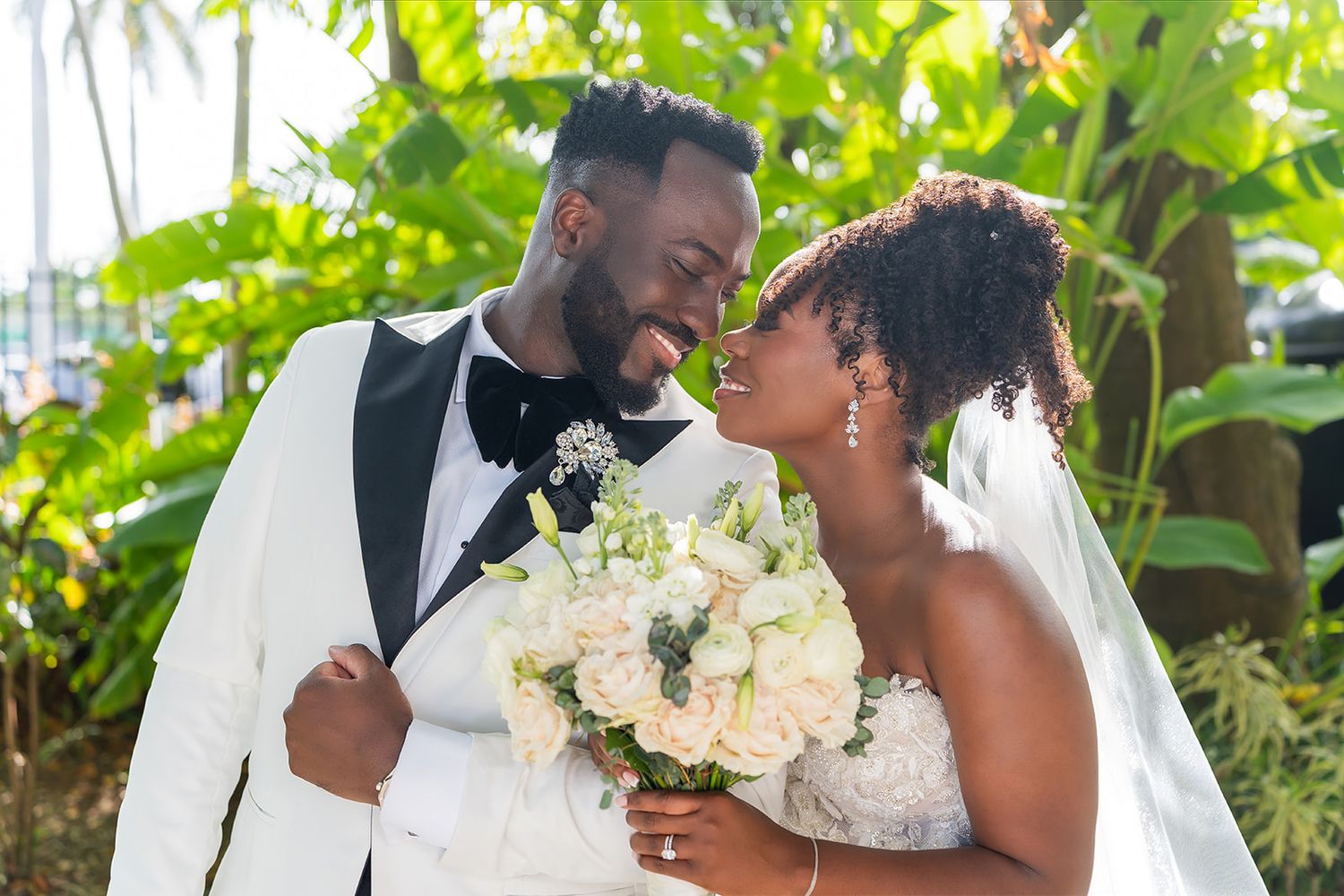 Joyful newlyweds share a romantic moment surrounded by tropical greenery holding white rose bouquet.