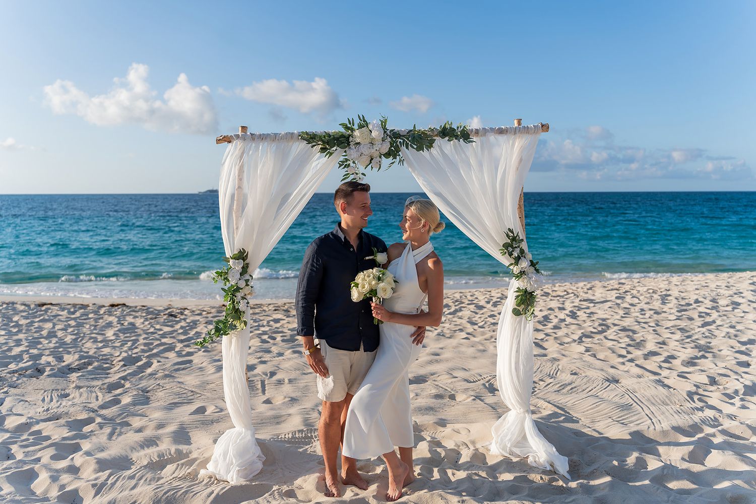 Couple posing under white draped wedding arch on tropical beach with turquoise ocean backdrop.