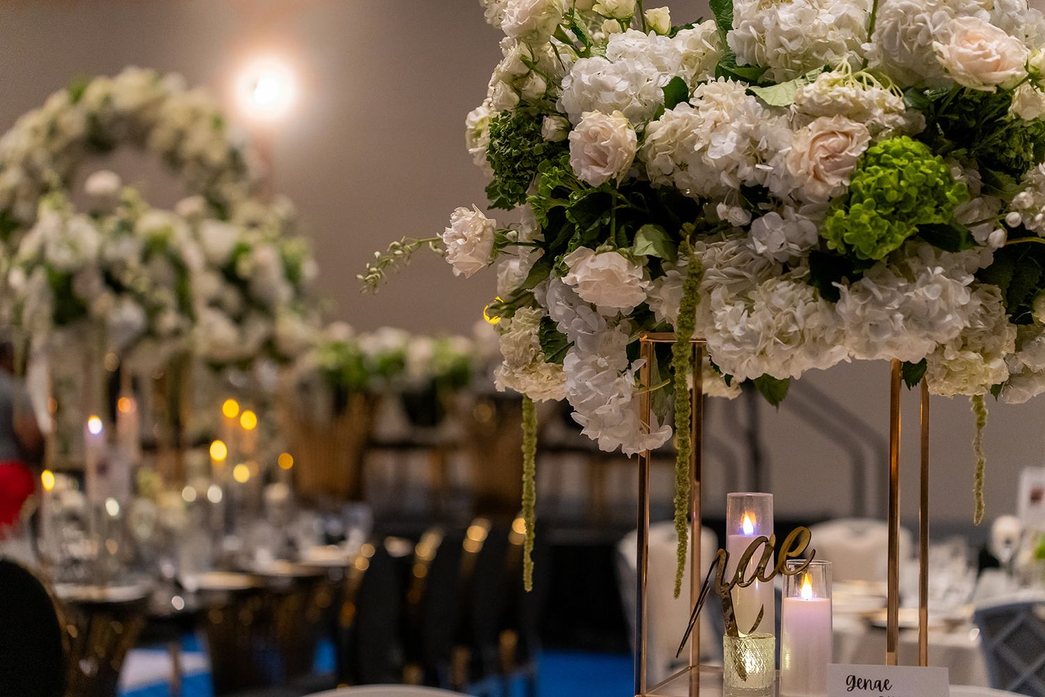 Elegant wedding reception centerpiece featuring white roses and green hydrangeas on tall gold stands with candles.