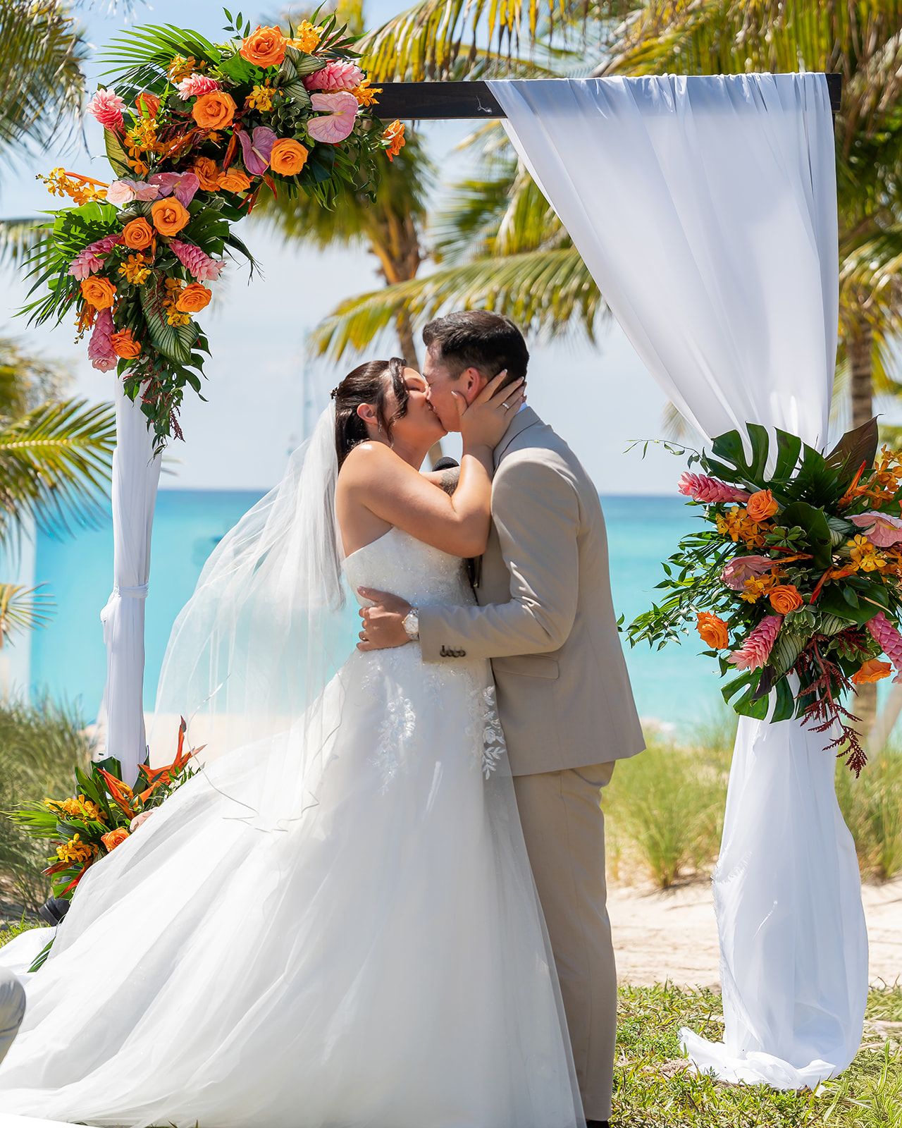 Bride and groom share first kiss under tropical floral arch with ocean backdrop at beach wedding ceremony.