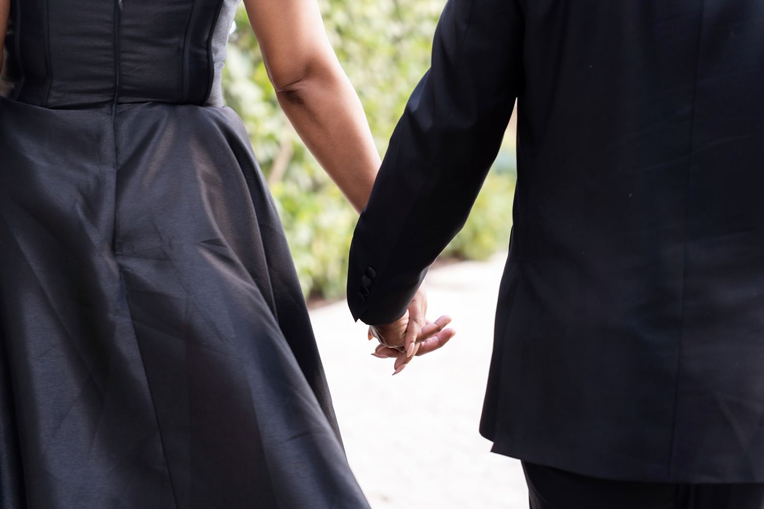 Couple holding hands in formal black attire, woman in elegant gown and man in suit, romantic outdoor setting.