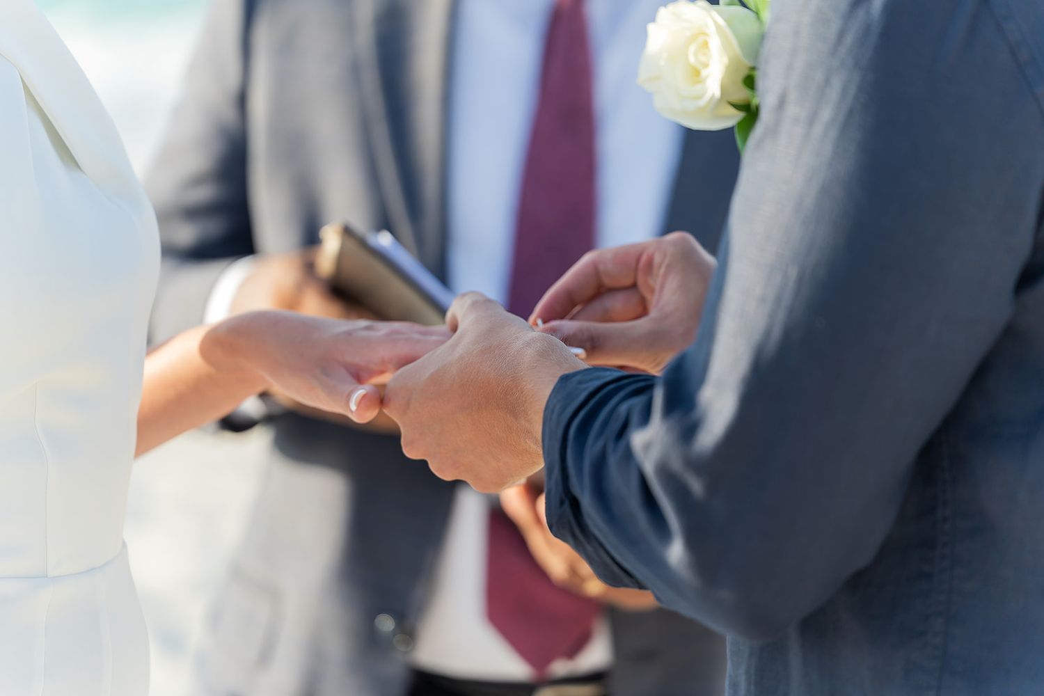 Groom placing wedding ring on bride's finger during outdoor beach ceremony, officiant holding book in background.