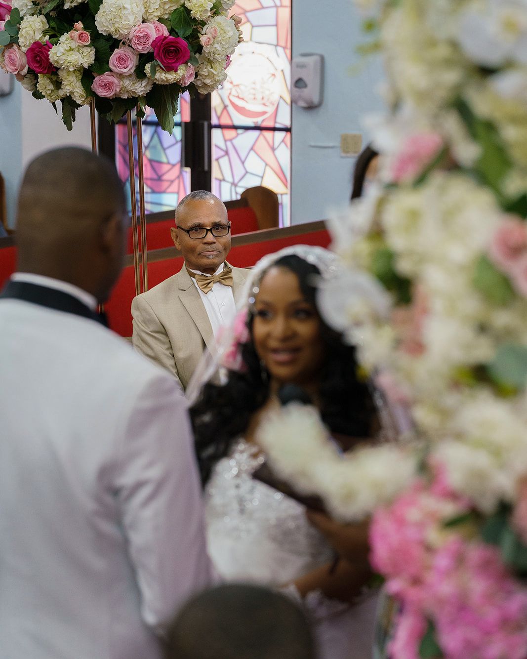 Groom in tan suit and bow tie watches bride walk down church aisle adorned with pink and white floral arrangements.