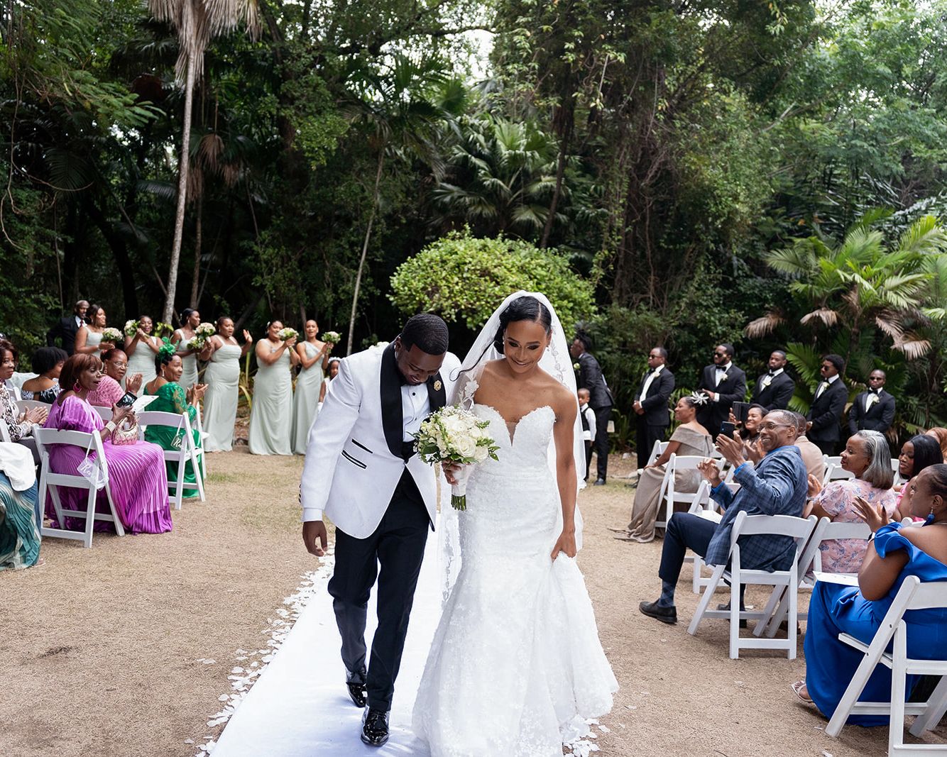 A bride and groom walk down the aisle at an outdoor wedding ceremony surrounded by guests and lush greenery.