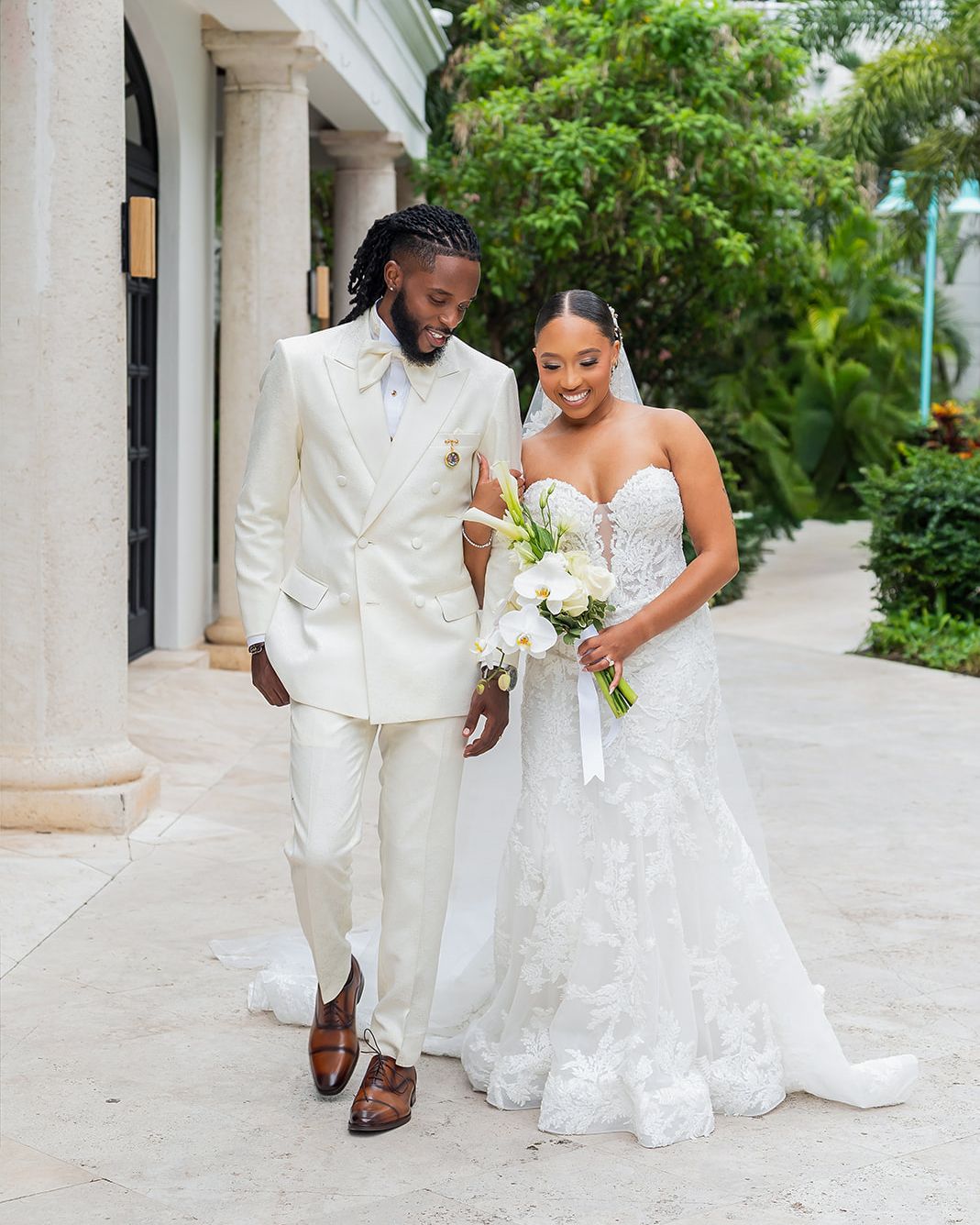 A bride and groom walk together outdoors, she in a lace gown holding flowers, he in a cream double-breasted suit.