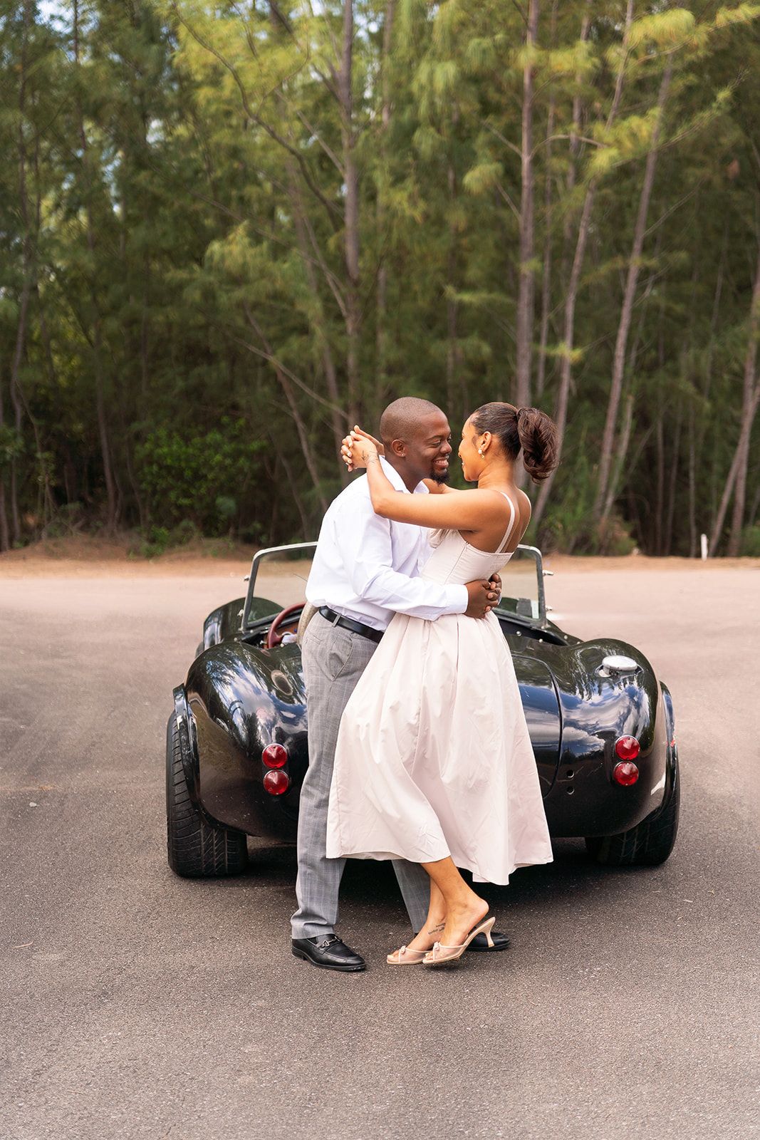 A couple embraces and dances beside a classic green convertible sports car in a scenic outdoor parking area.