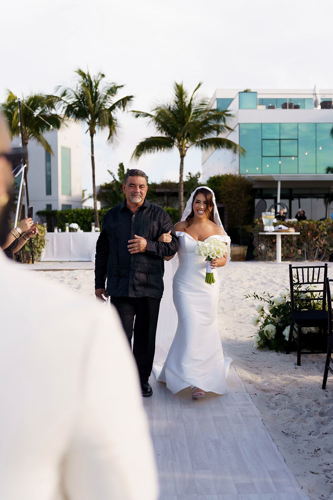 Bride in white gown walks down sandy beach aisle with escort near palm trees at outdoor wedding ceremony.