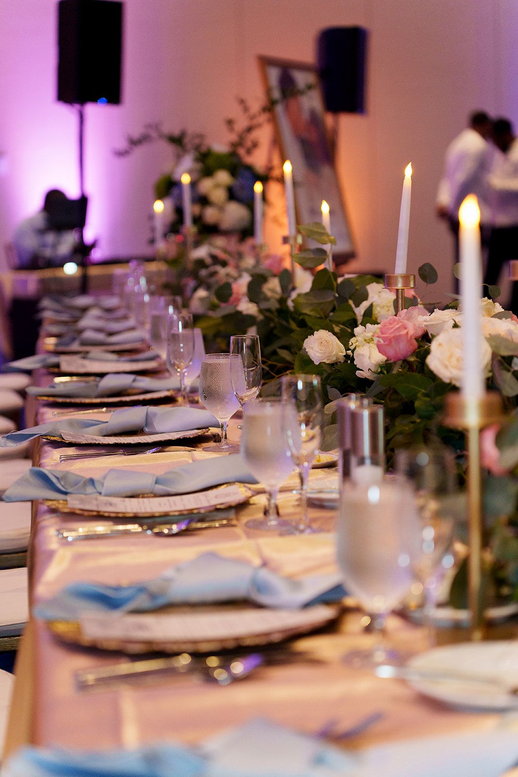 Elegant wedding reception table with pink roses, candles, gold chargers, and blue napkins in a softly lit ballroom.