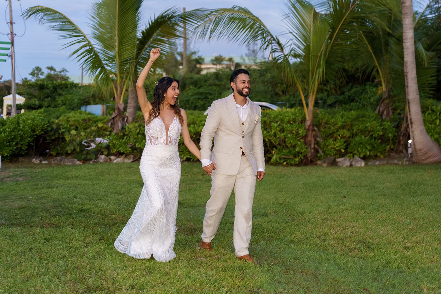 Bride in white gown and groom in tan suit hold hands celebrating outdoors on tropical lawn with palm trees.