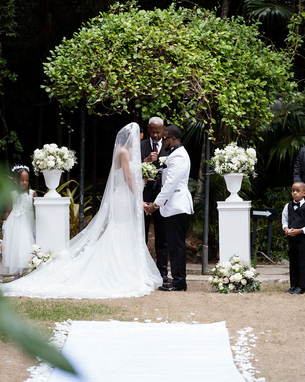 Outdoor wedding ceremony with bride in white gown and veil, groom in white jacket, officiant, flower girl, and ring bearer.