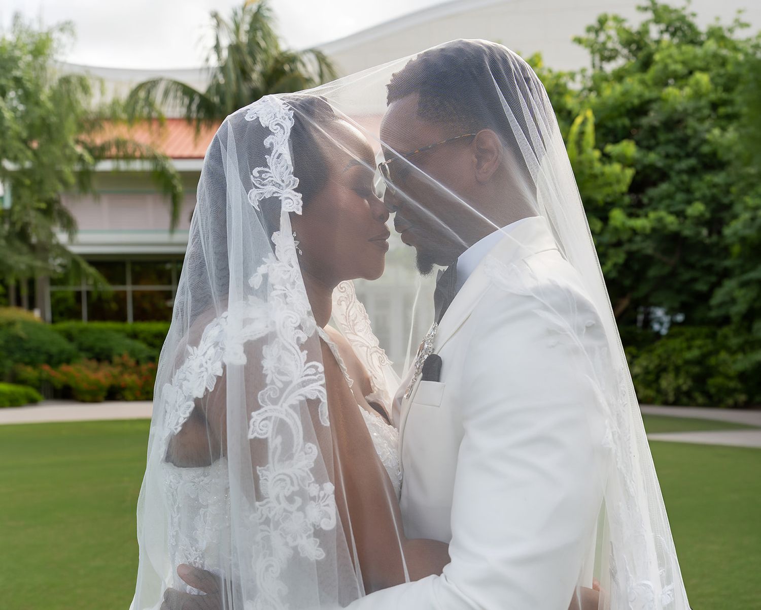 A bride and groom share a romantic kiss under a flowing white lace wedding veil outdoors on their wedding day.