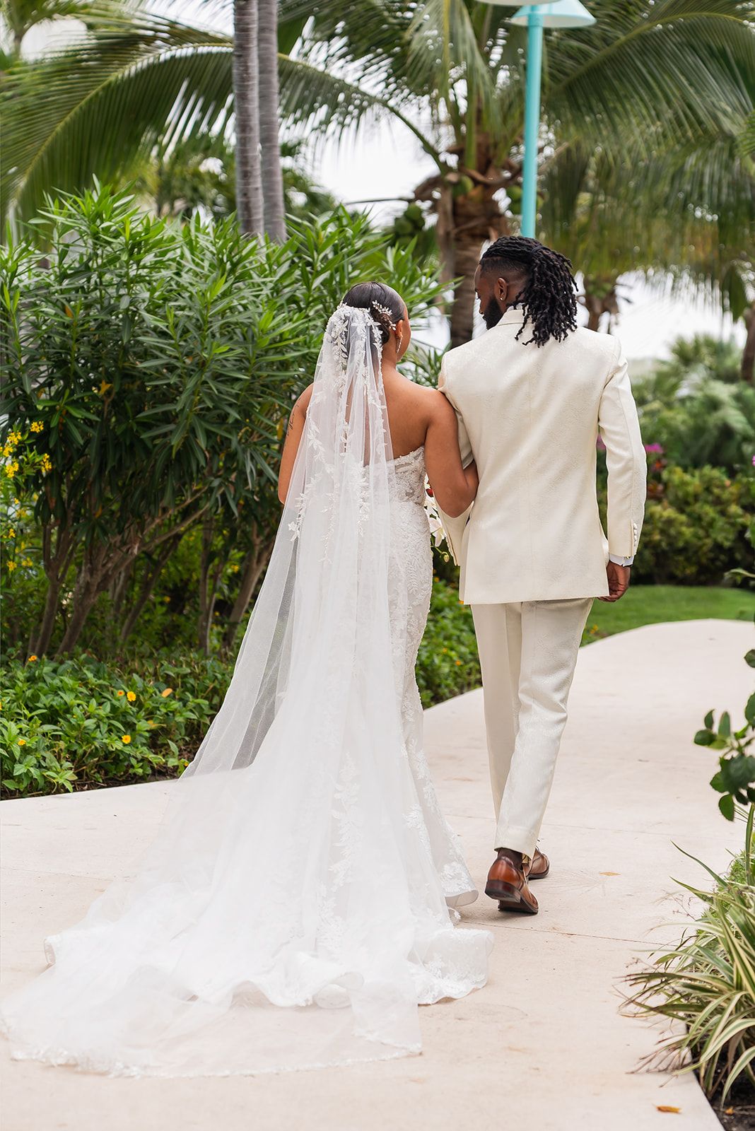 A bride and groom walk hand-in-hand along a tropical path, her lace gown and veil trailing behind them.