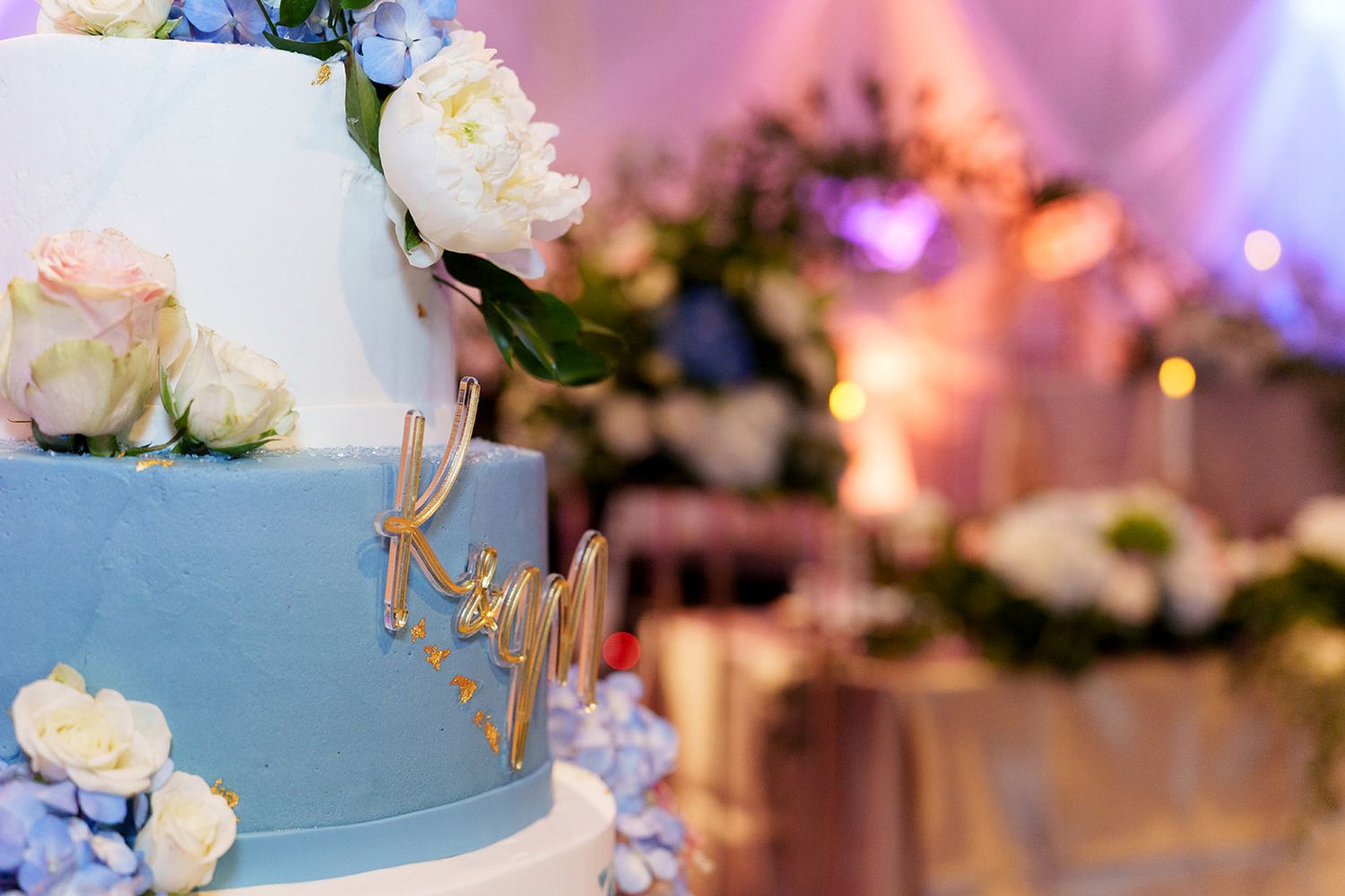 Elegant two-tier blue and white wedding cake with gold name script and fresh white roses and hydrangeas.