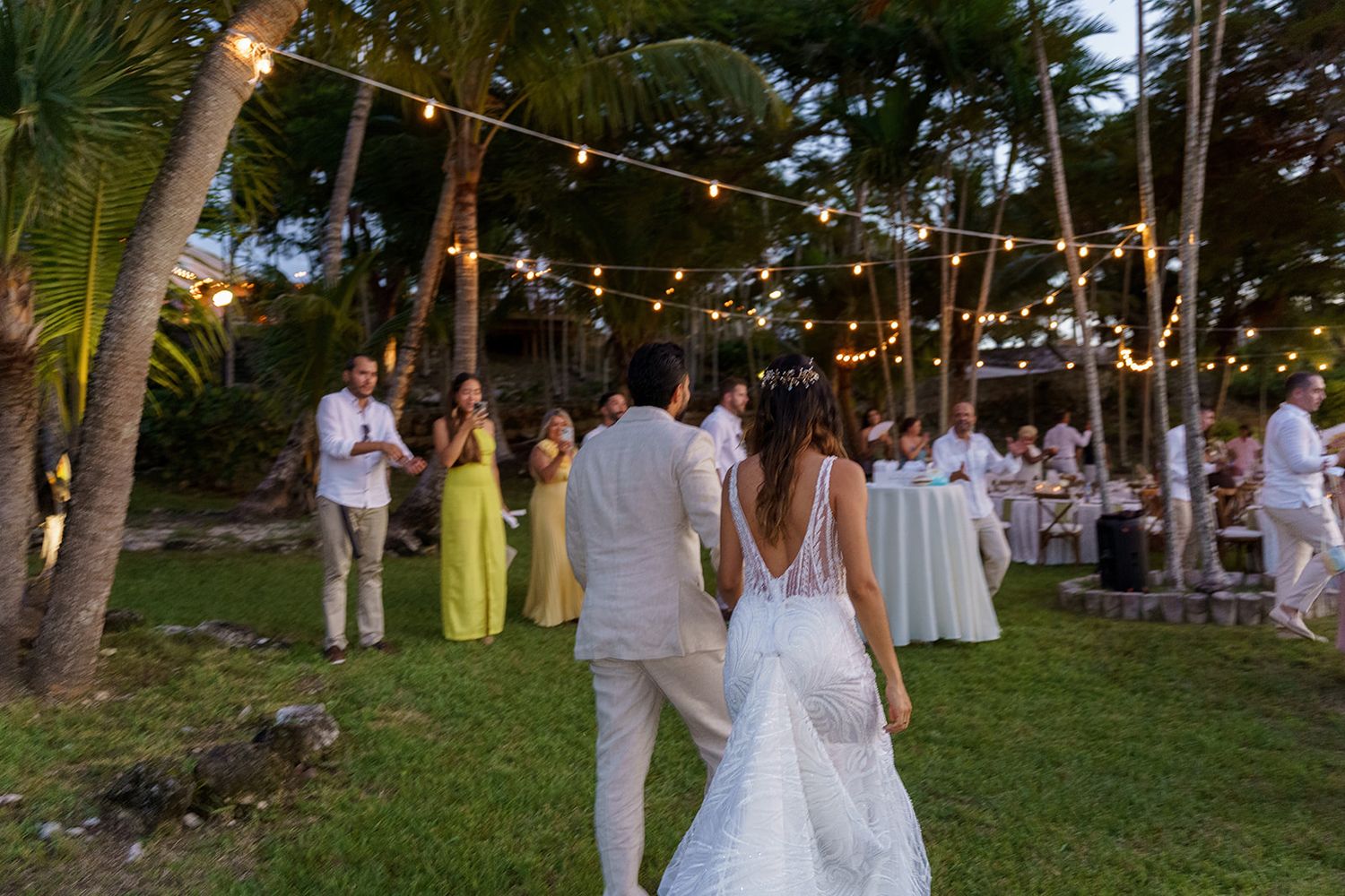 Couple walks hand-in-hand at outdoor wedding reception with string lights, palm trees, and guests in white and yellow attire.