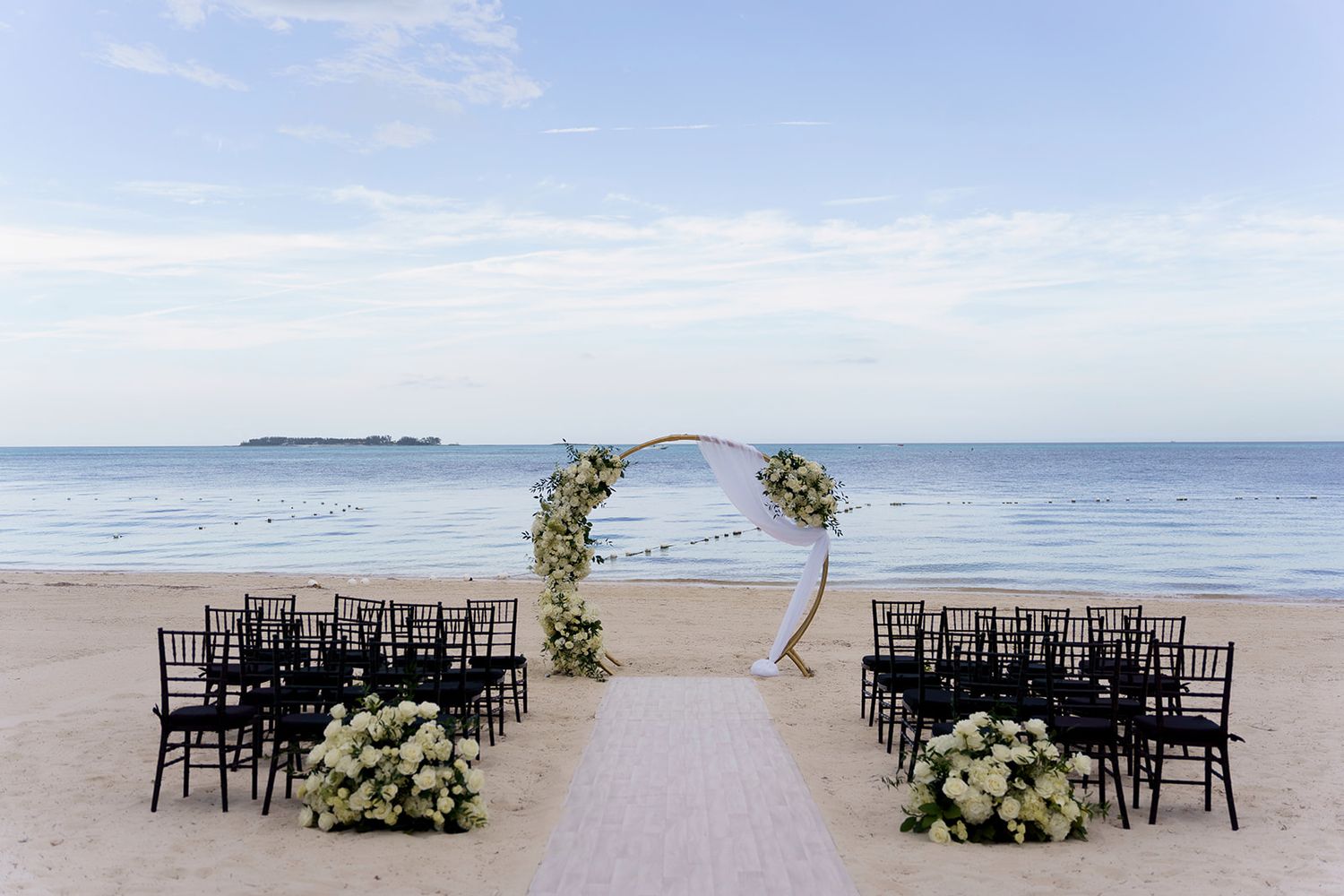 Elegant beach wedding setup with floral arch, black chairs, and white flowers by the ocean.
