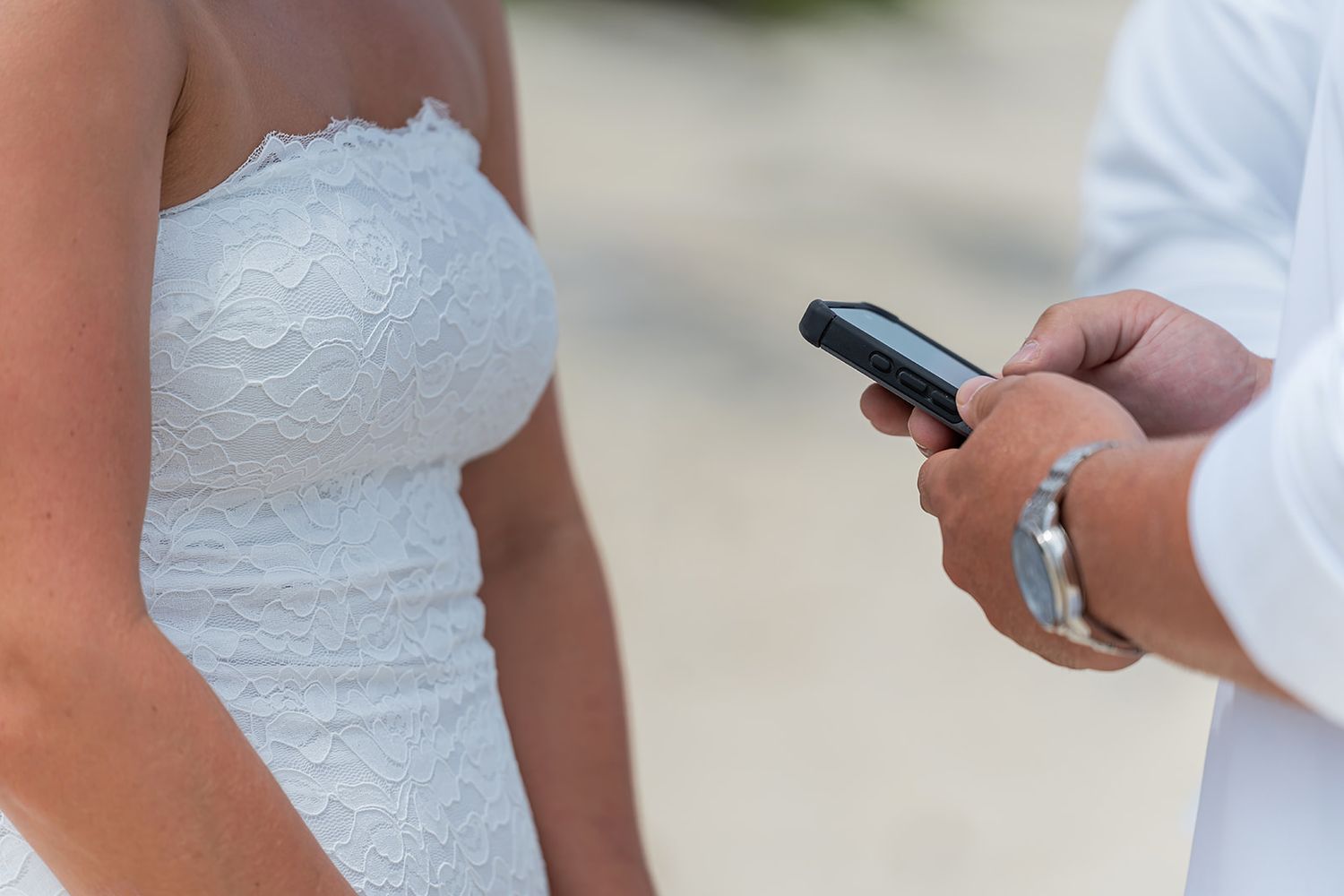 Person in white wedding dress stands next to someone in white shirt using a black smartphone outdoors.