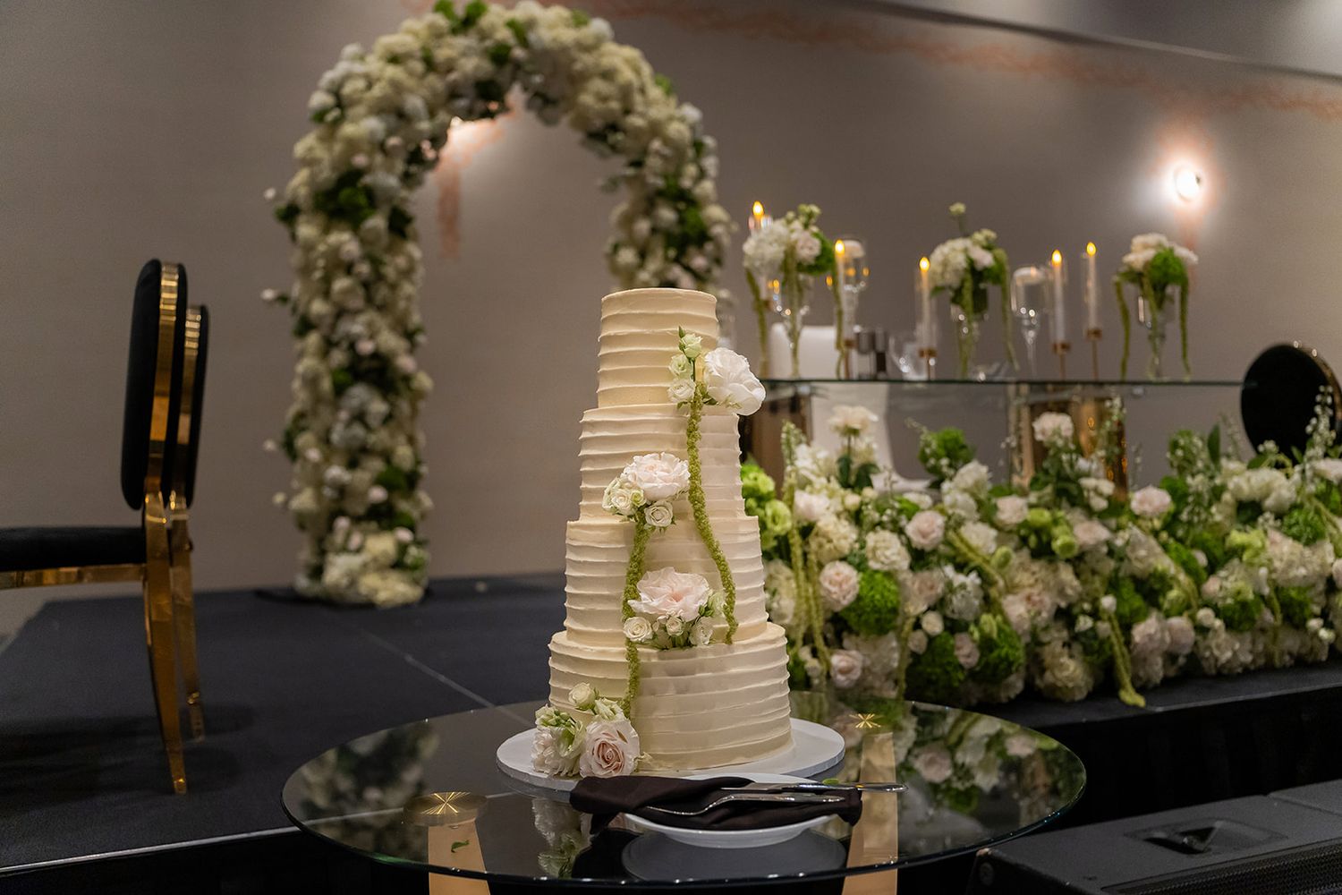 Elegant multi-tiered white wedding cake adorned with roses, set against a floral arch backdrop with candles at a reception.