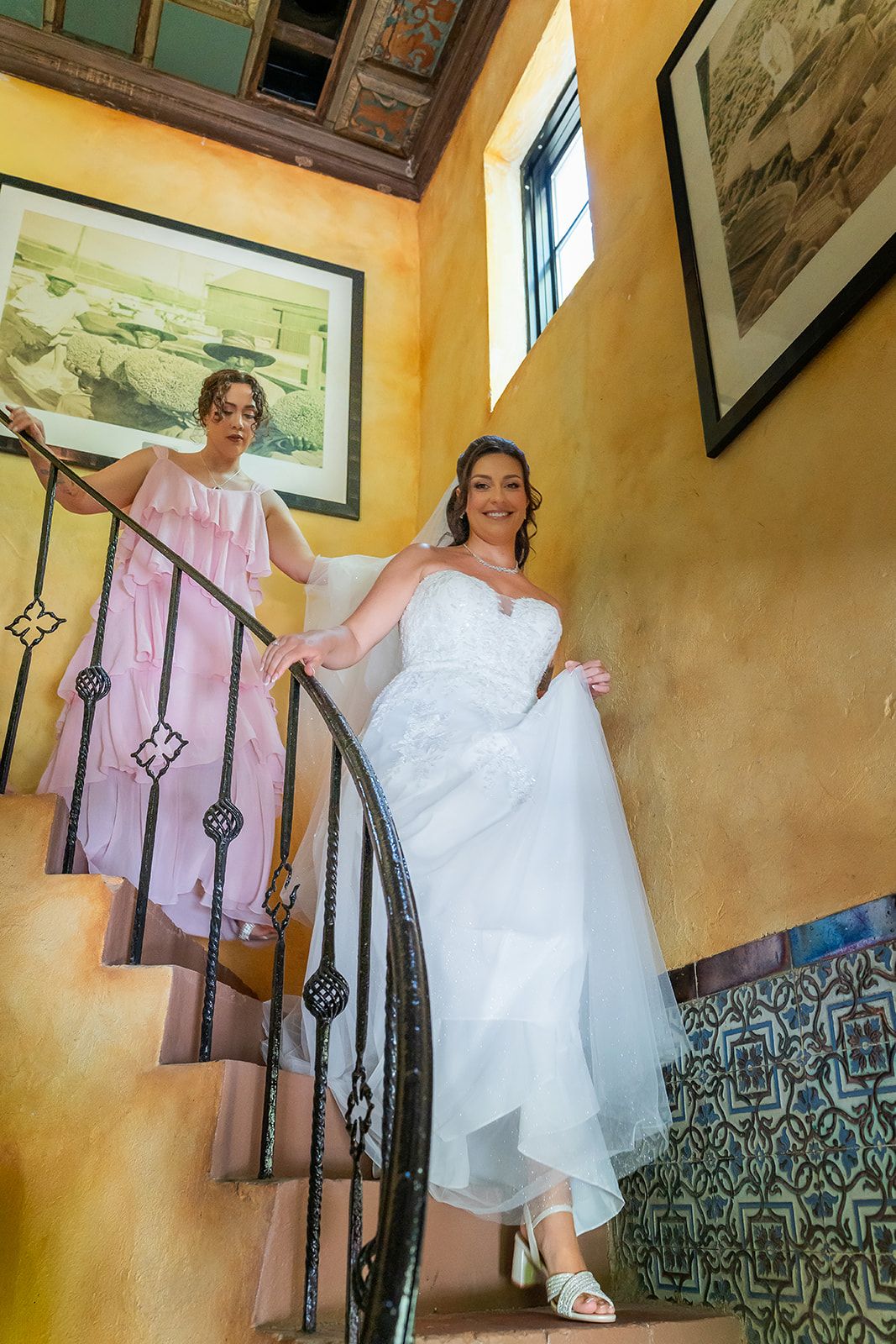 Bride in white gown descends ornate staircase assisted by bridesmaid in pink dress at elegant wedding venue.