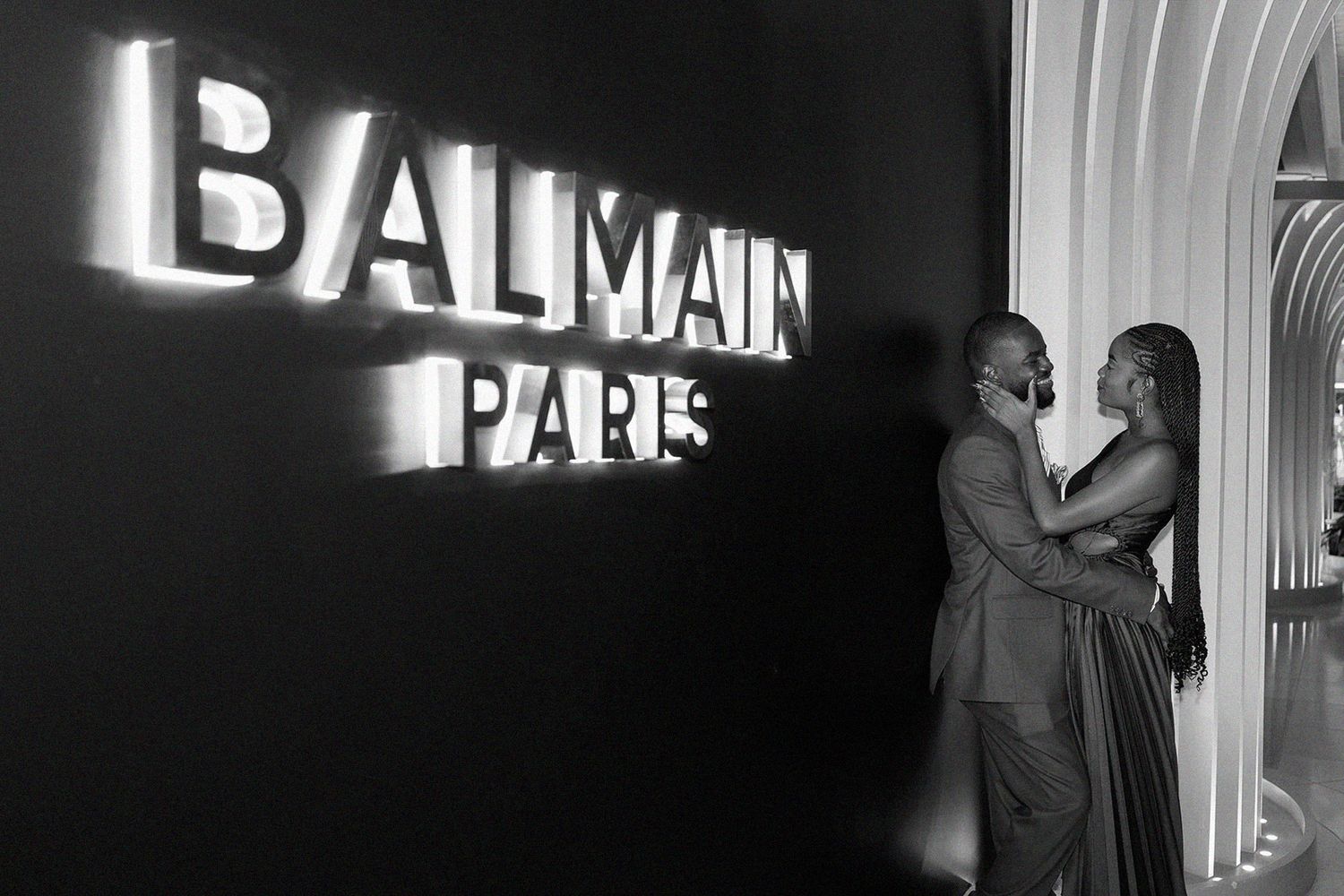 Couple embracing in black and white photo in front of illuminated Balmain Paris sign at luxury fashion event.