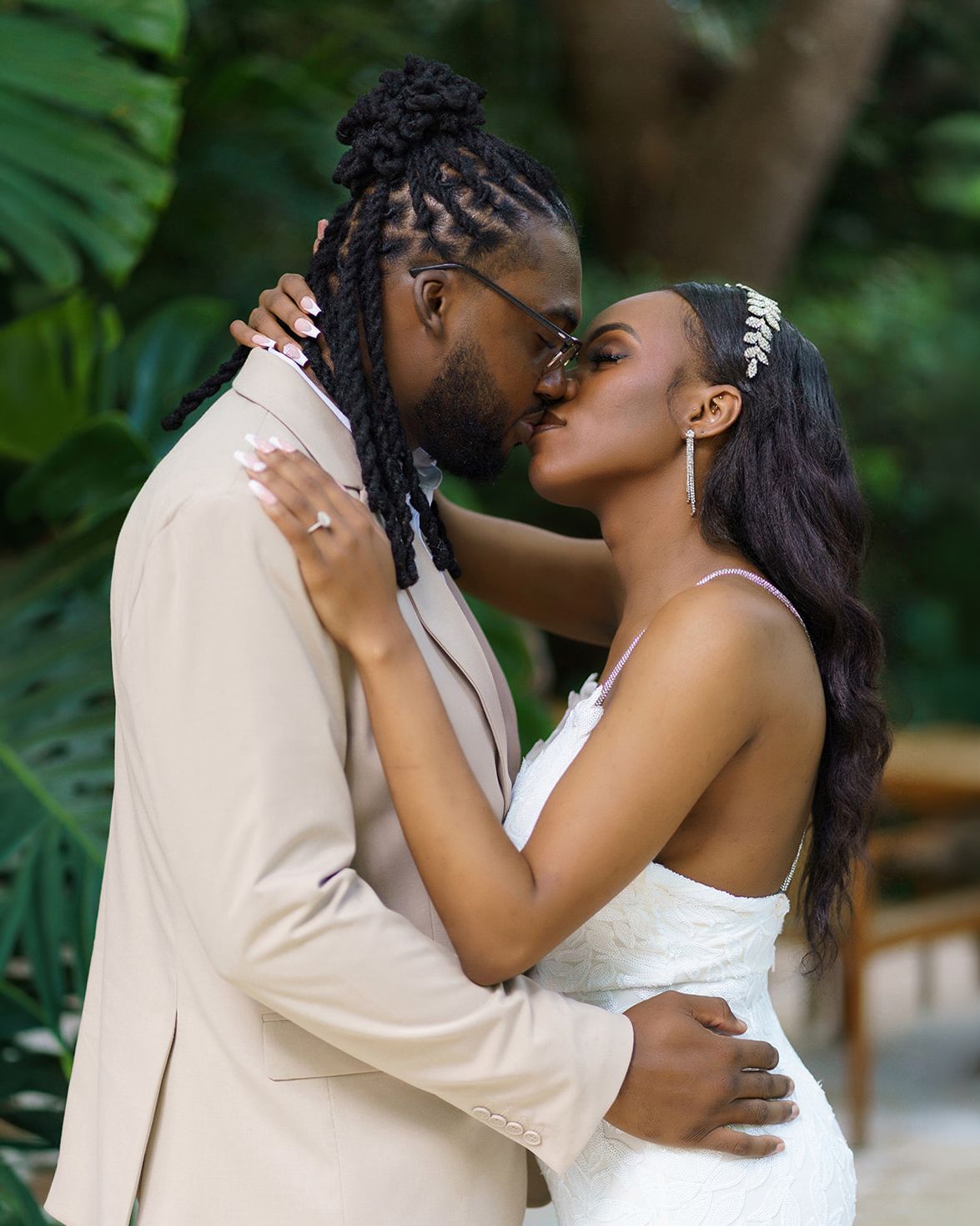 A couple shares a romantic kiss outdoors, dressed in wedding attire, surrounded by lush tropical greenery.