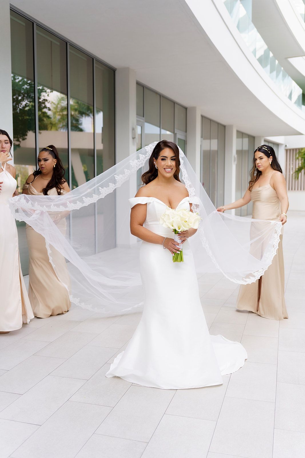 Bride in white off-shoulder gown holds bouquet while bridesmaids in champagne dresses spread her lace veil outdoors.