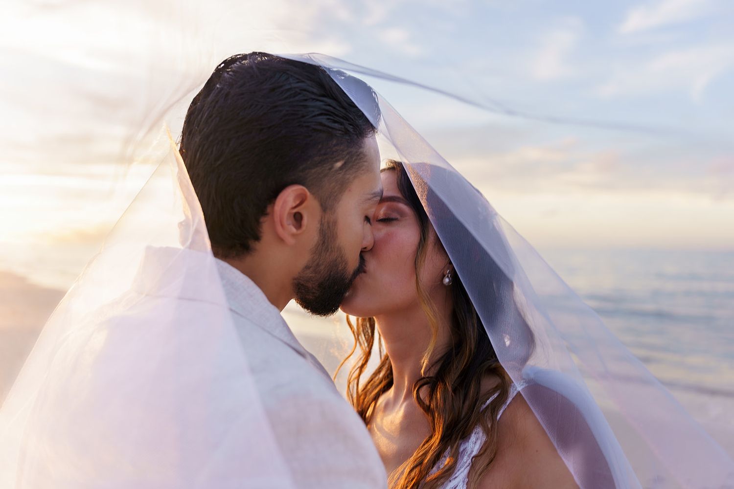 Bride and groom sharing a romantic kiss under a wedding veil on a beach at sunset.