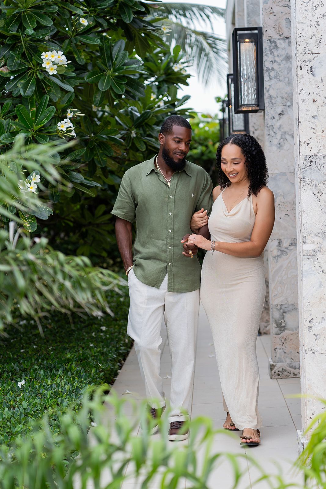 A couple walks together along a garden pathway, man in green shirt and white pants, woman in beige dress.