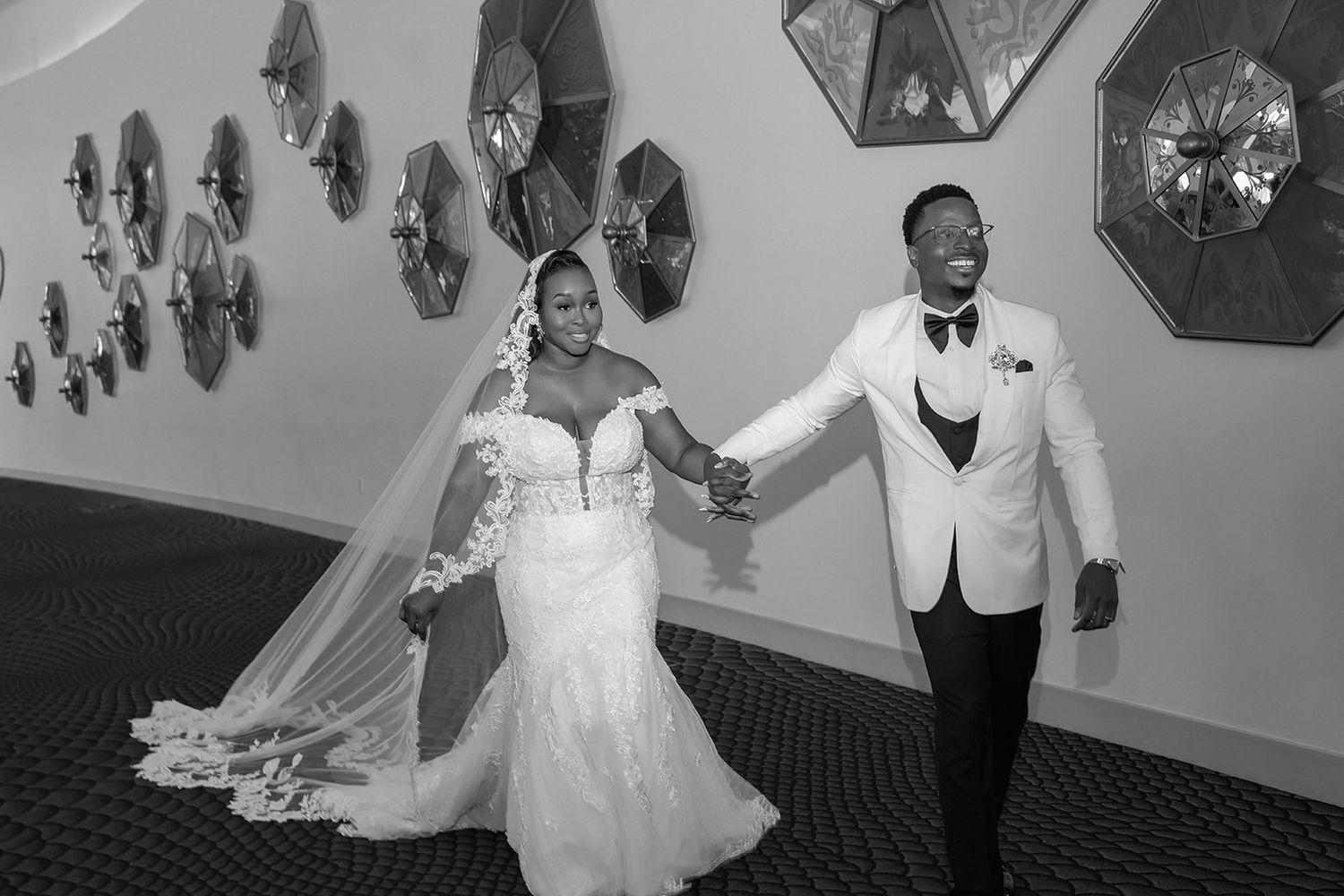 Black and white photo of a bride and groom holding hands and walking together in a decorated wedding venue.