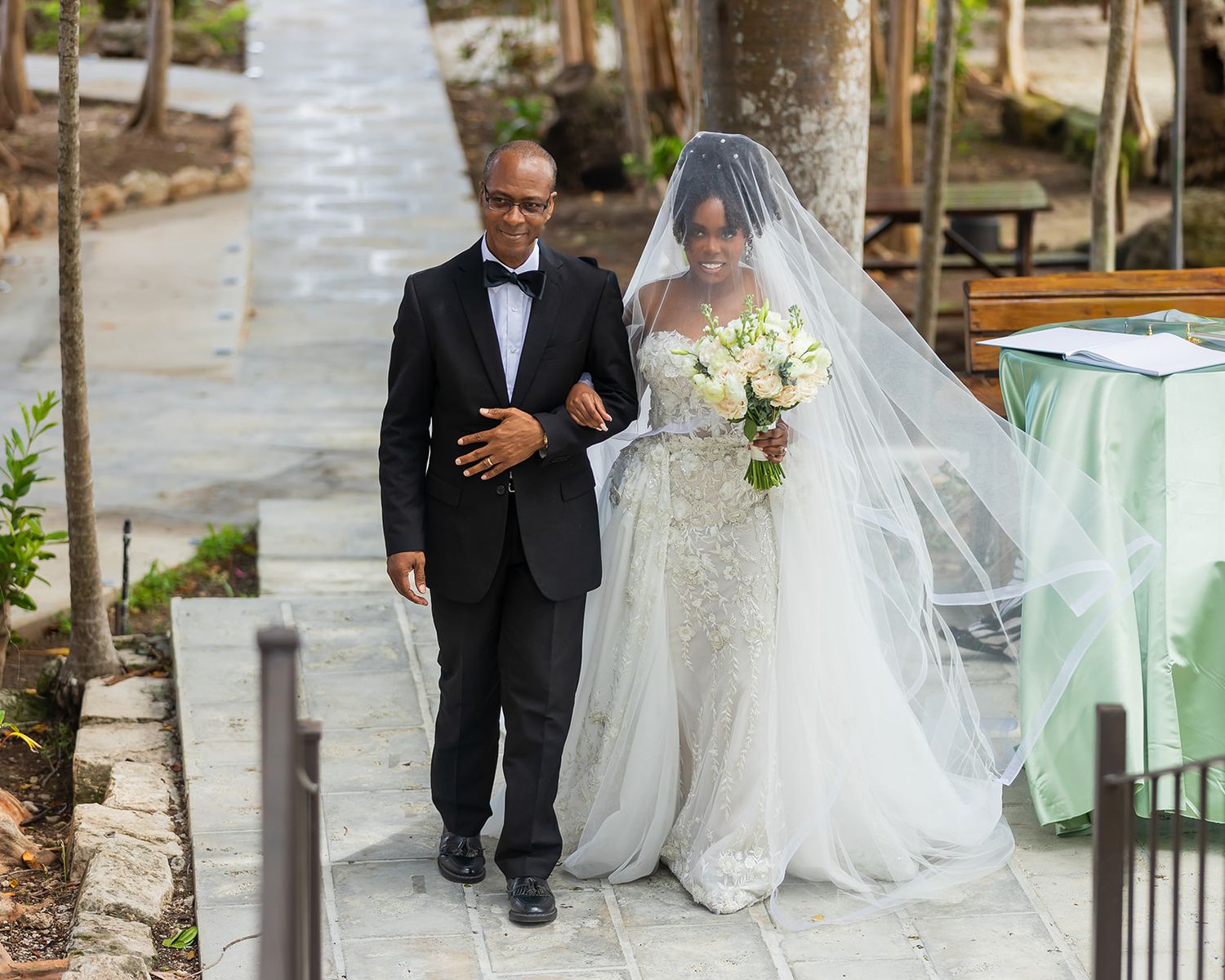 Father escorts bride in stunning lace gown and long veil down the outdoor wedding aisle.