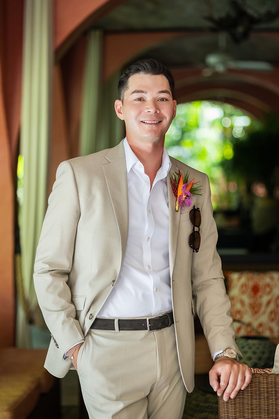 Smiling groom in beige suit with tropical boutonniere and sunglasses, posing in elegant outdoor venue.