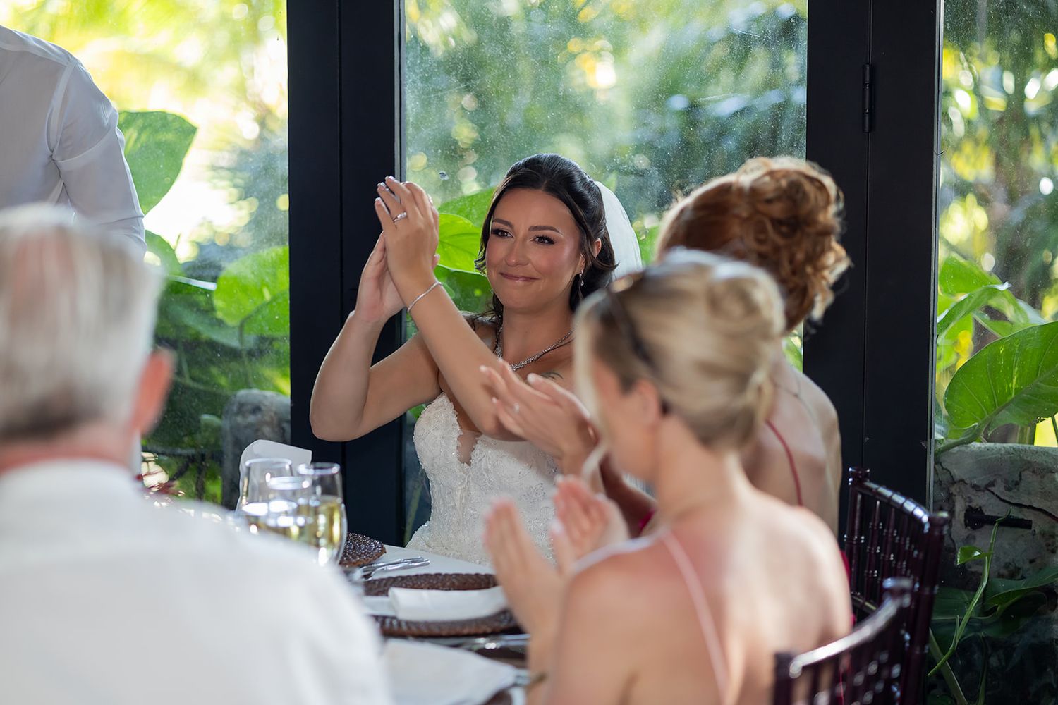 Bride clapping and smiling with guests at wedding reception table, surrounded by lush greenery.