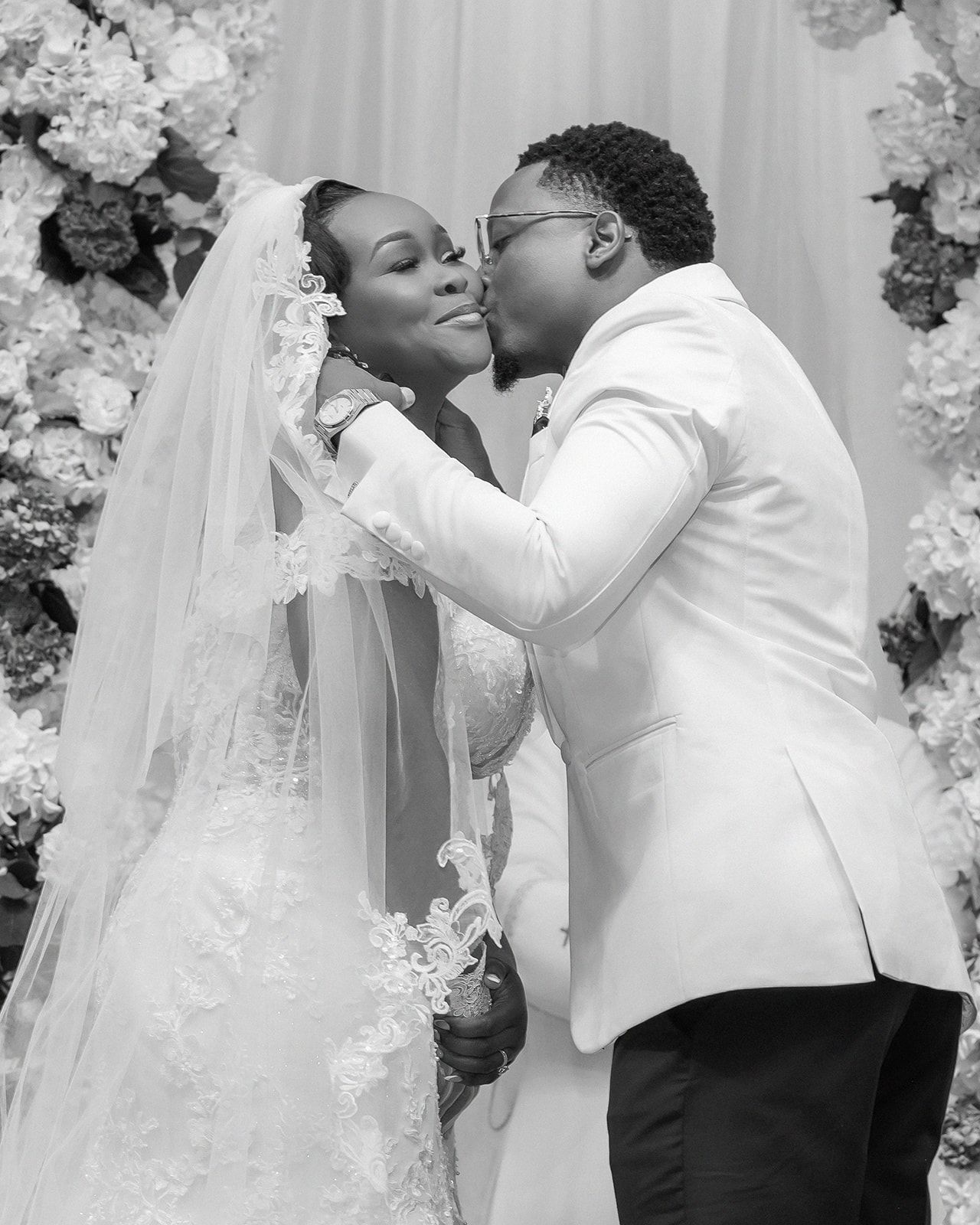 Black and white wedding photo of a groom in white tuxedo kissing bride in veil before floral backdrop.