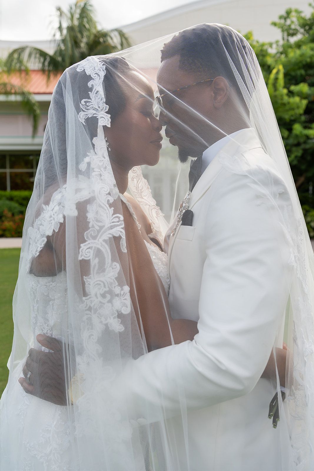 A bride and groom share an intimate moment under a lace wedding veil outdoors, dressed in white wedding attire.