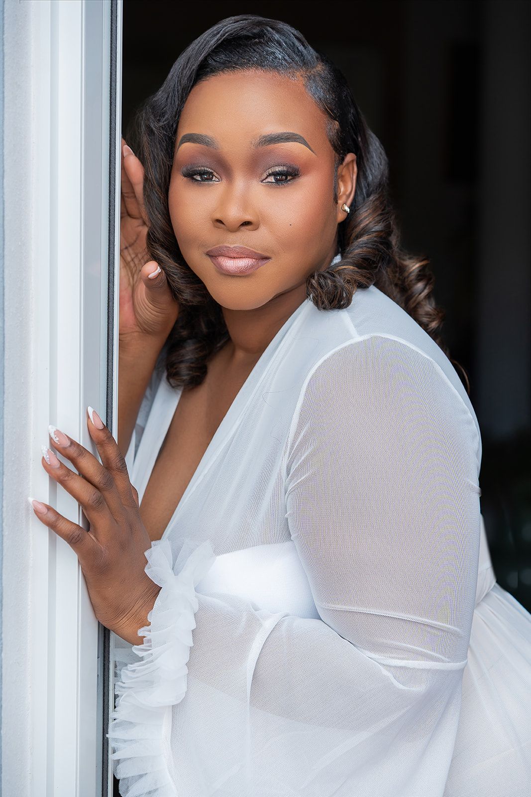 A woman in a white sheer dress poses elegantly by a doorframe with styled curls and glam makeup.