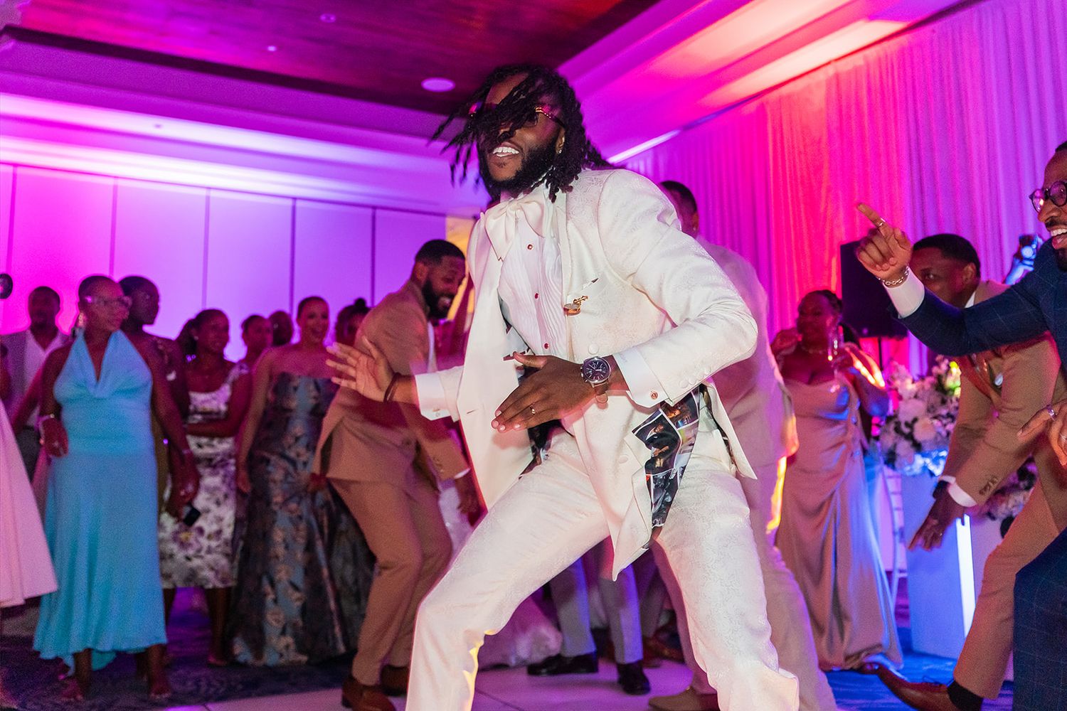 Man in white suit dancing energetically at a wedding reception with guests celebrating in the background.