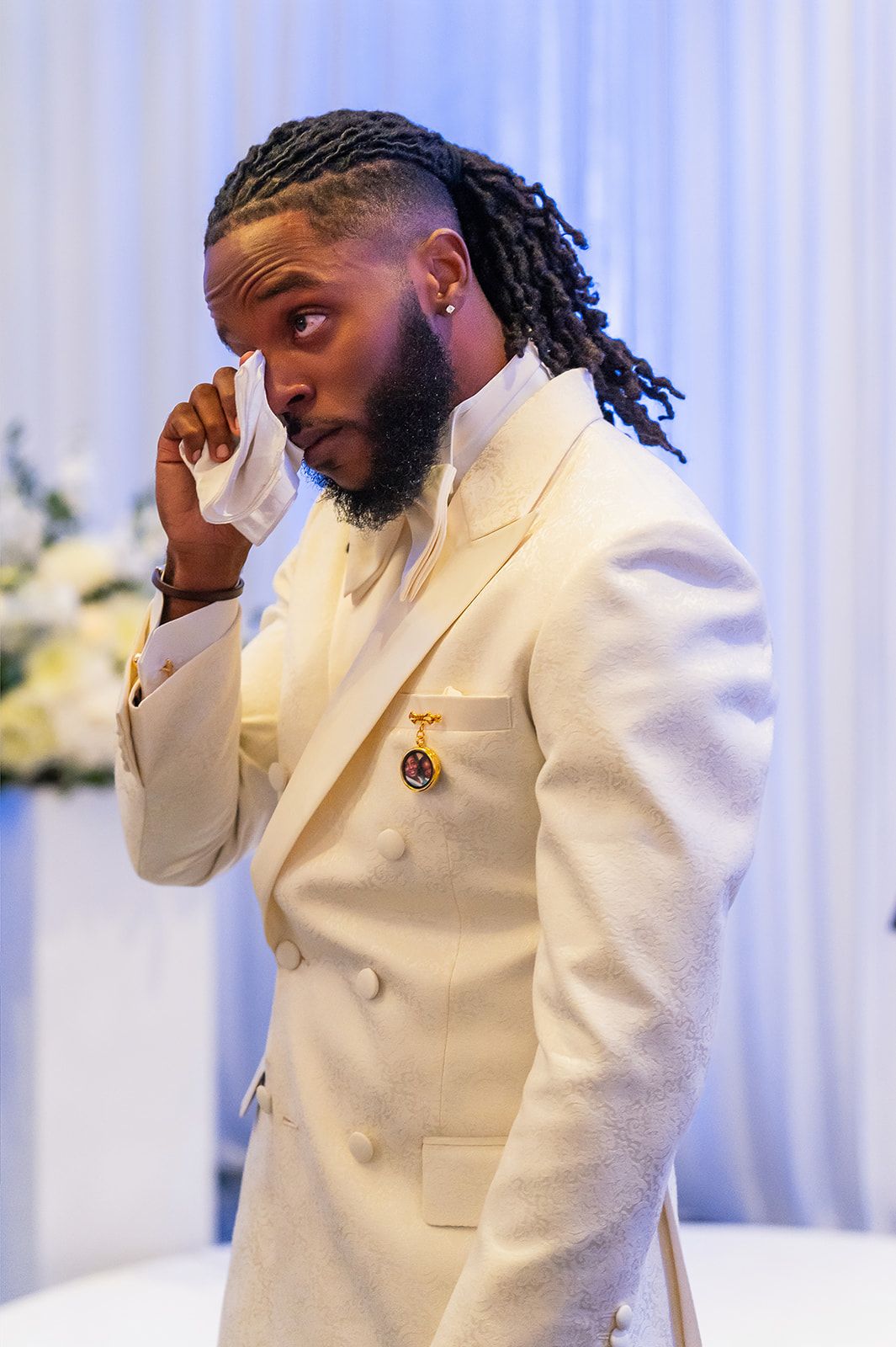 Emotional man in white tuxedo wiping tear at formal event.