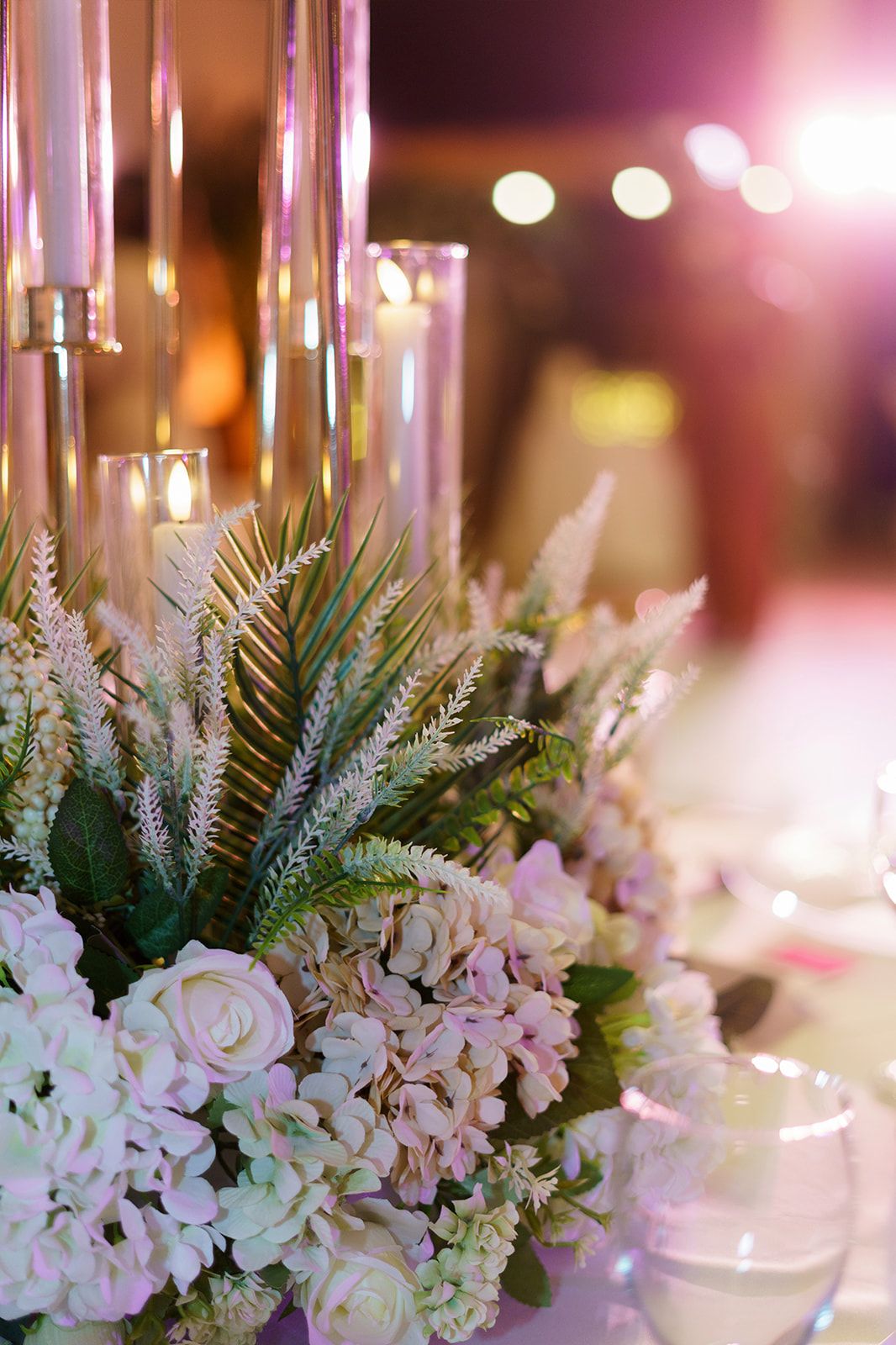 Elegant wedding table centerpiece with white roses, hydrangeas, greenery, and tall glass candle holders glowing softly.