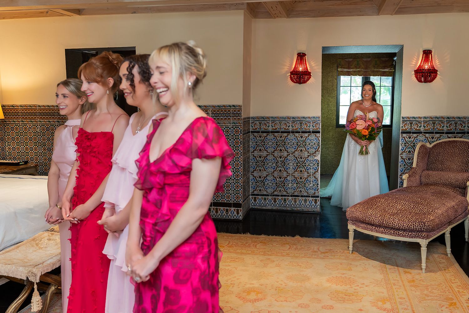 Bridesmaids in pink dresses stand together as bride holding bouquet waits in doorway during wedding preparations.