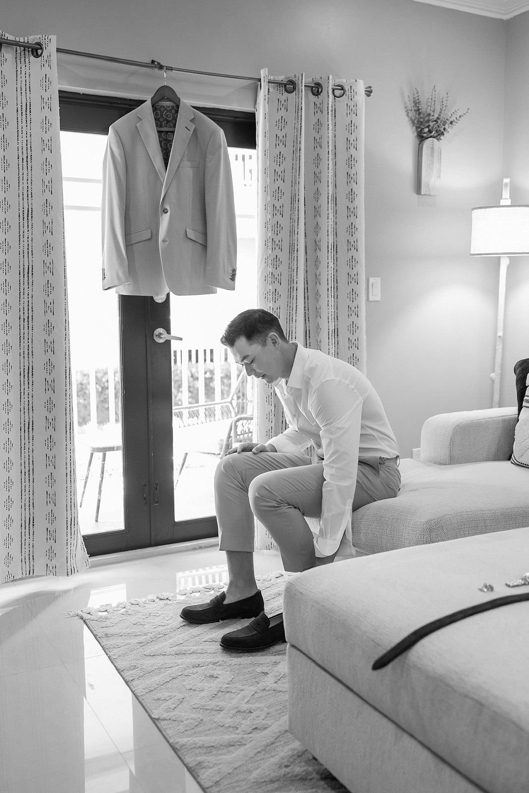 Groom sitting on couch putting on shoes in black and white photo with suit jacket hanging on door.