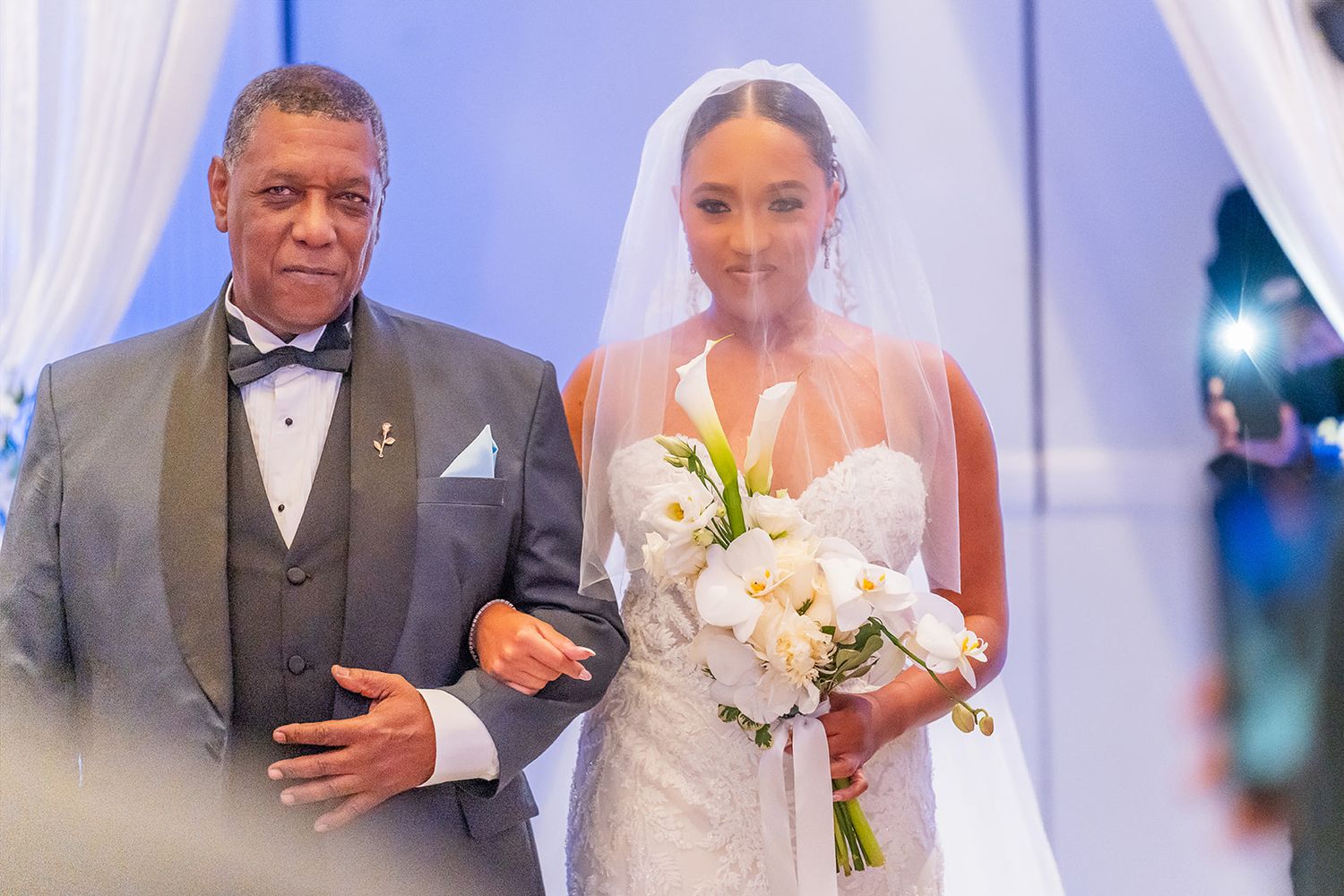 Bride walks down aisle with escort holding white floral bouquet at wedding ceremony.