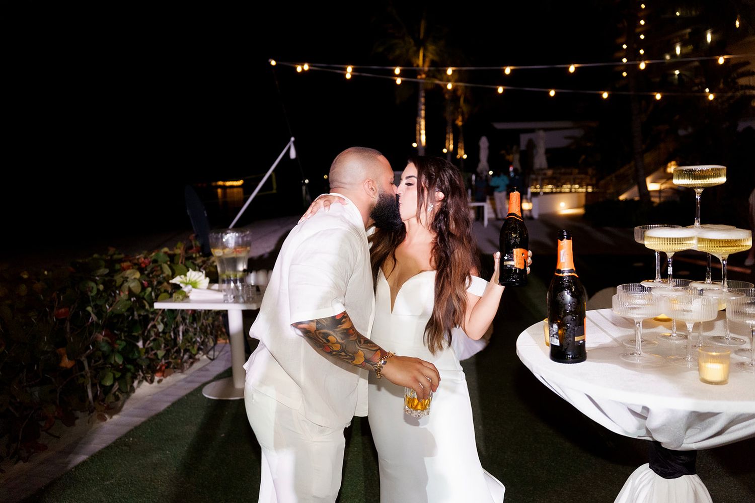 Bride and groom share a kiss at outdoor wedding reception at night with champagne tower and string lights.