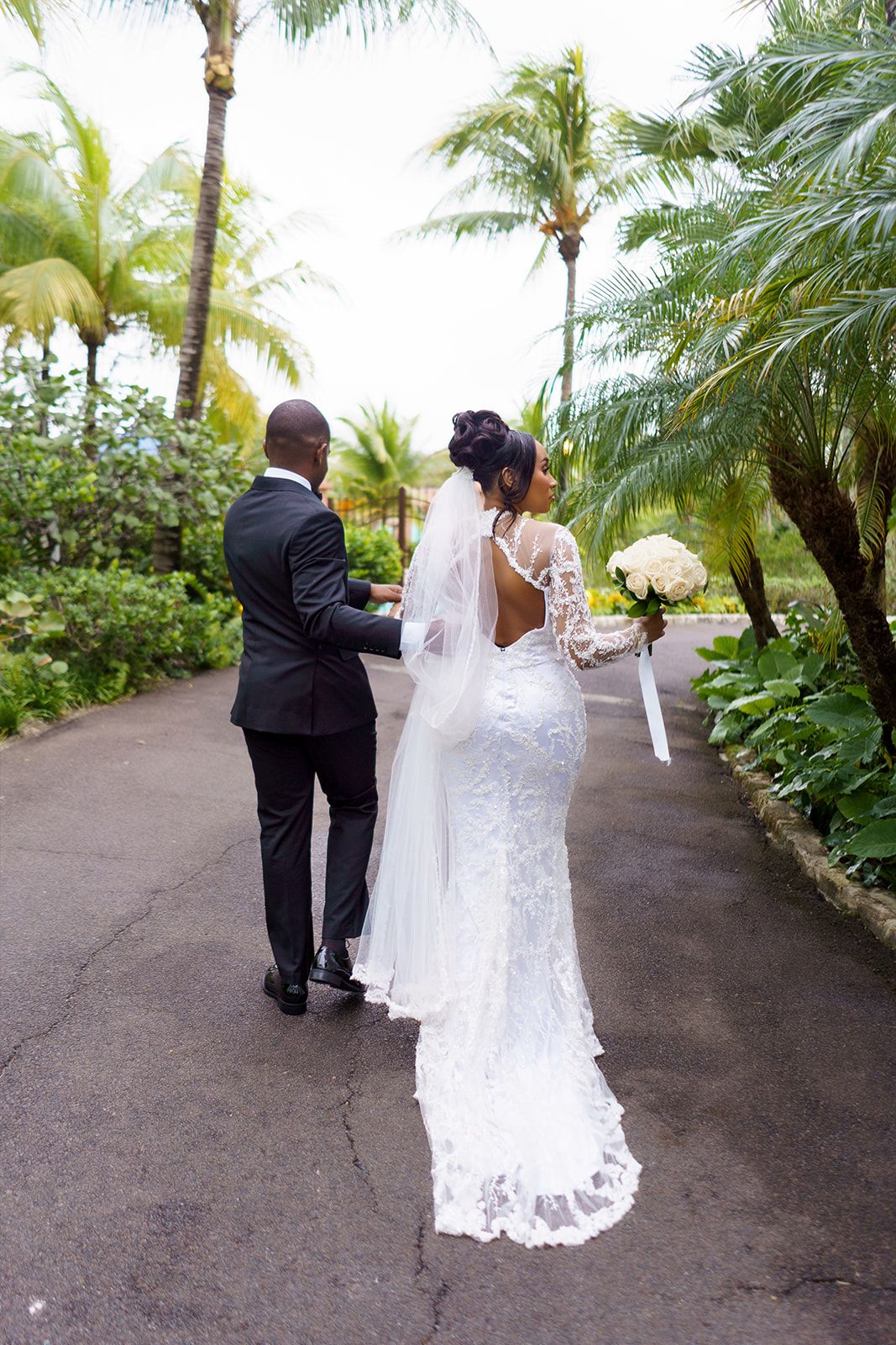 Bride and groom walking hand in hand down a tropical palm-lined path, bride wearing a lace backless wedding gown with veil.