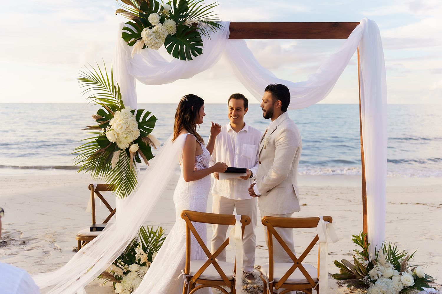 Couple exchanges vows under decorated wooden arch on beach with white draping and tropical floral arrangements.